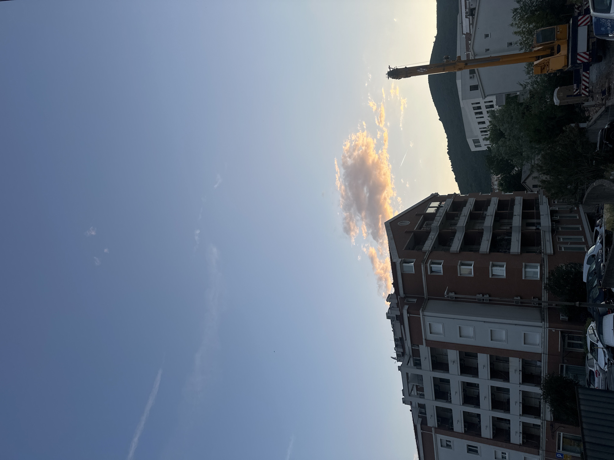 The image is an upward-facing shot of the sky and surrounding cityscape of Budva, Montenegro. The sky is predominantly blue with a small, peach-colored cloud near the center, appearing to be lit by the setting sun. A thin contrail is visible to the cloud's right. In the top-right corner, there is a crane with a yellow boom arm. Behind the crane is a hill with vegetation and a white building with multiple windows. In the bottom-right, a multi-story residential building with red brick and white sections, balconies, and windows, is visible. Parked cars and trees appear at the base of the building, with a glimpse of a police car also noticeable.