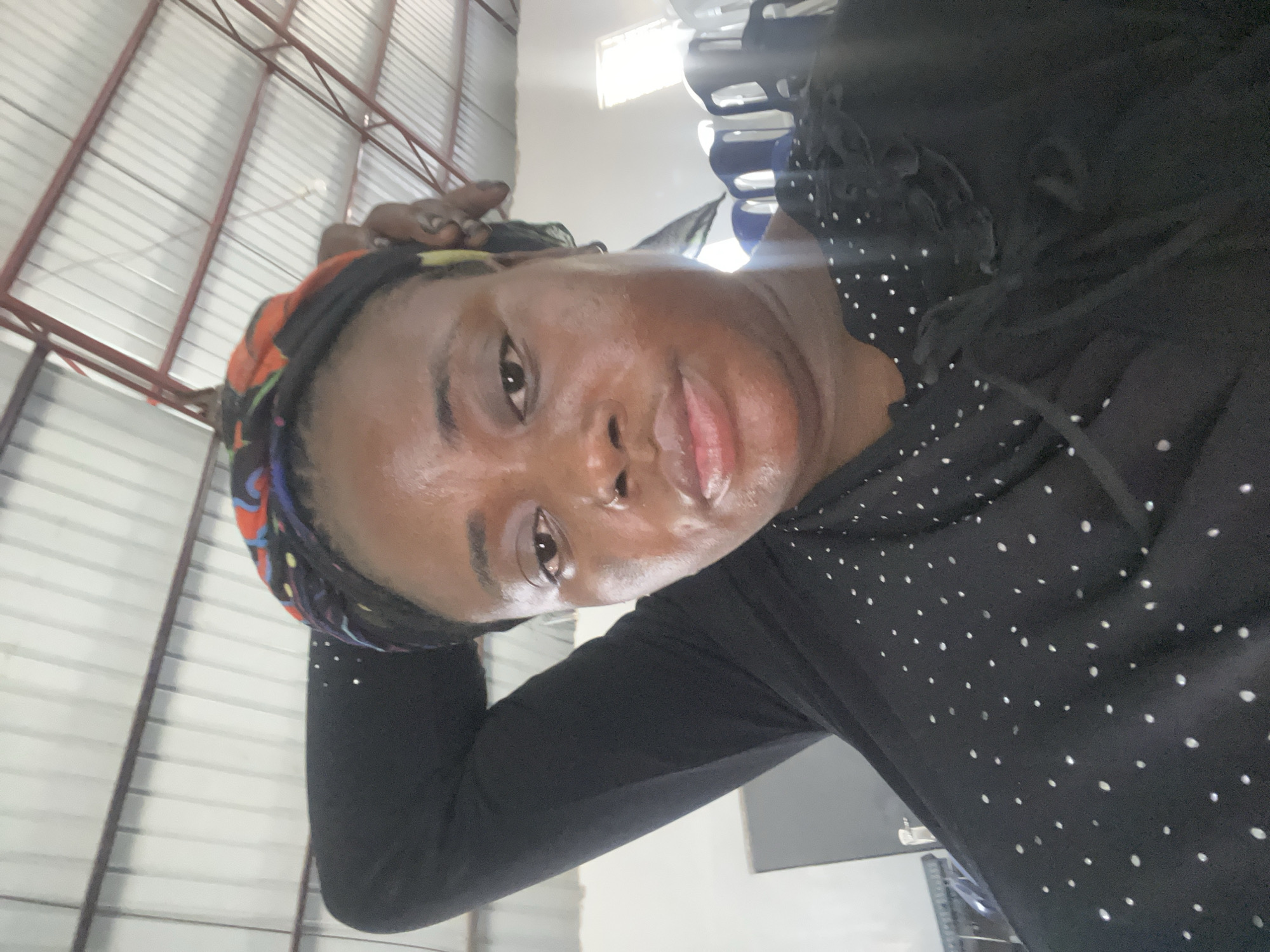 A close-up, tilted selfie of a dark-skinned woman with a patterned headscarf, wearing a black, long-sleeved top with white dots. She is indoors under a corrugated metal ceiling with exposed red beams. Blue and white chairs are visible in the background. The photo appears to have been taken in Barakin Akawo, Nigeria.