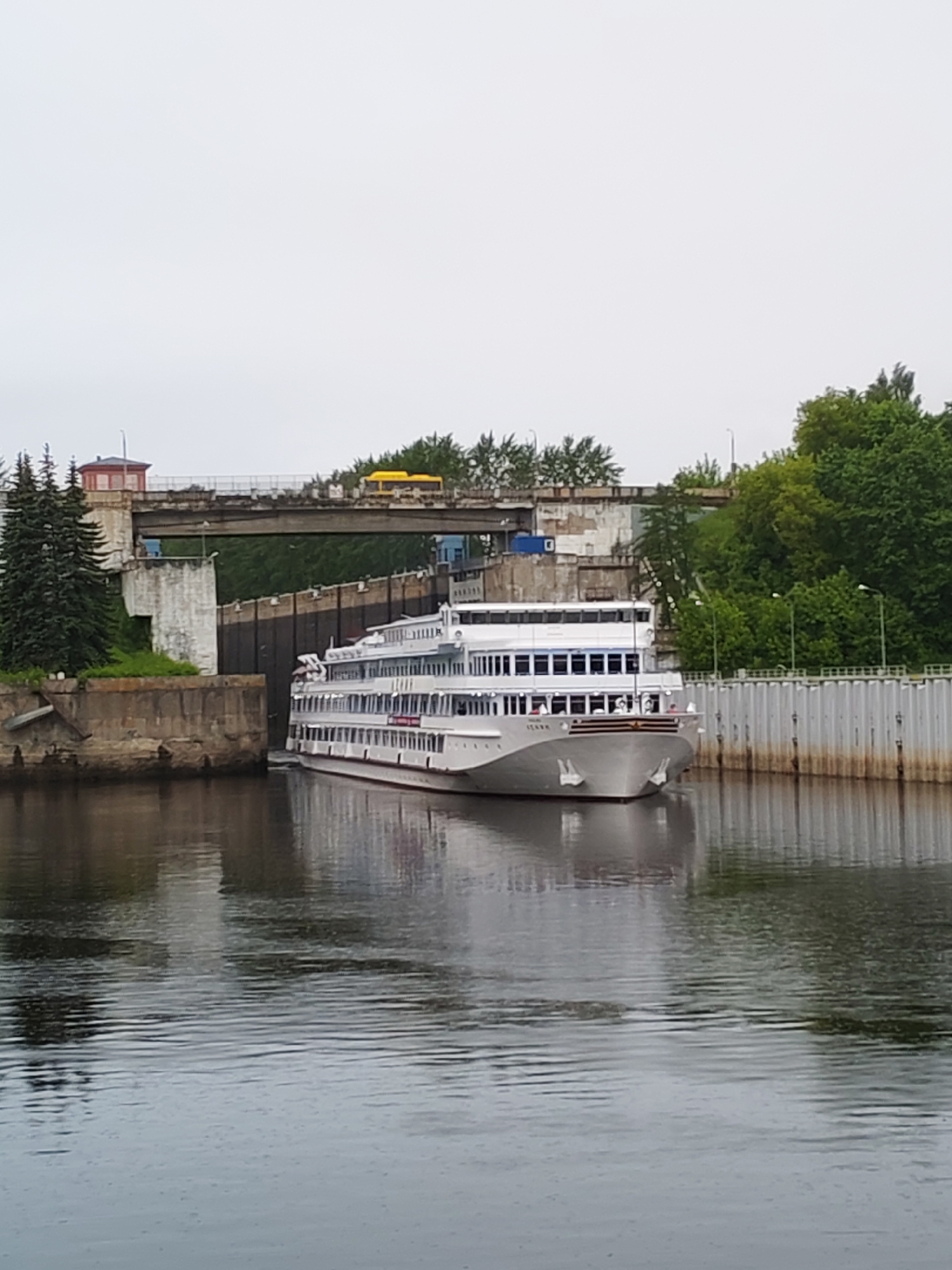 A large, white, multi-decked river cruise ship is positioned in the lock system of a canal in Bol'shie Mkhi, Russia. The ship is partially obscured by the concrete walls of the lock. Above the lock, a concrete bridge spans the canal, and a small, red-roofed structure sits to the left of the bridge. A yellow vehicle is also visible on the bridge. Green trees and other vegetation line the banks of the canal. The water is dark and reflective, showing the ship and surrounding structures. The sky is overcast and grey. A metal barrier is located on the right side of the canal.