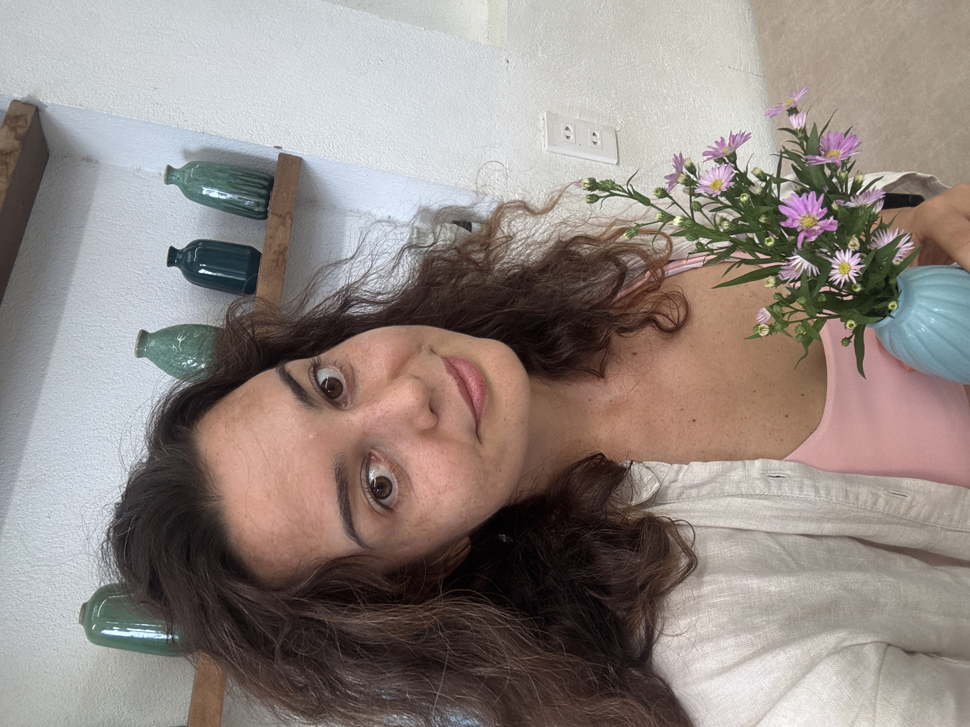 A woman with curly brown hair and fair skin, wearing a pink top and a light-colored unbuttoned shirt, holds a small blue vase filled with light purple flowers. She is positioned in the foreground, looking towards the camera with a slight smile. In the background, a white wall is visible, with a white electrical outlet on it. Above, a wooden shelf holds several green ceramic vases of varying shapes and sizes. The overall setting appears to be indoors, possibly a room or a cafe in Hoi An, Vietnam, given the decorative elements.