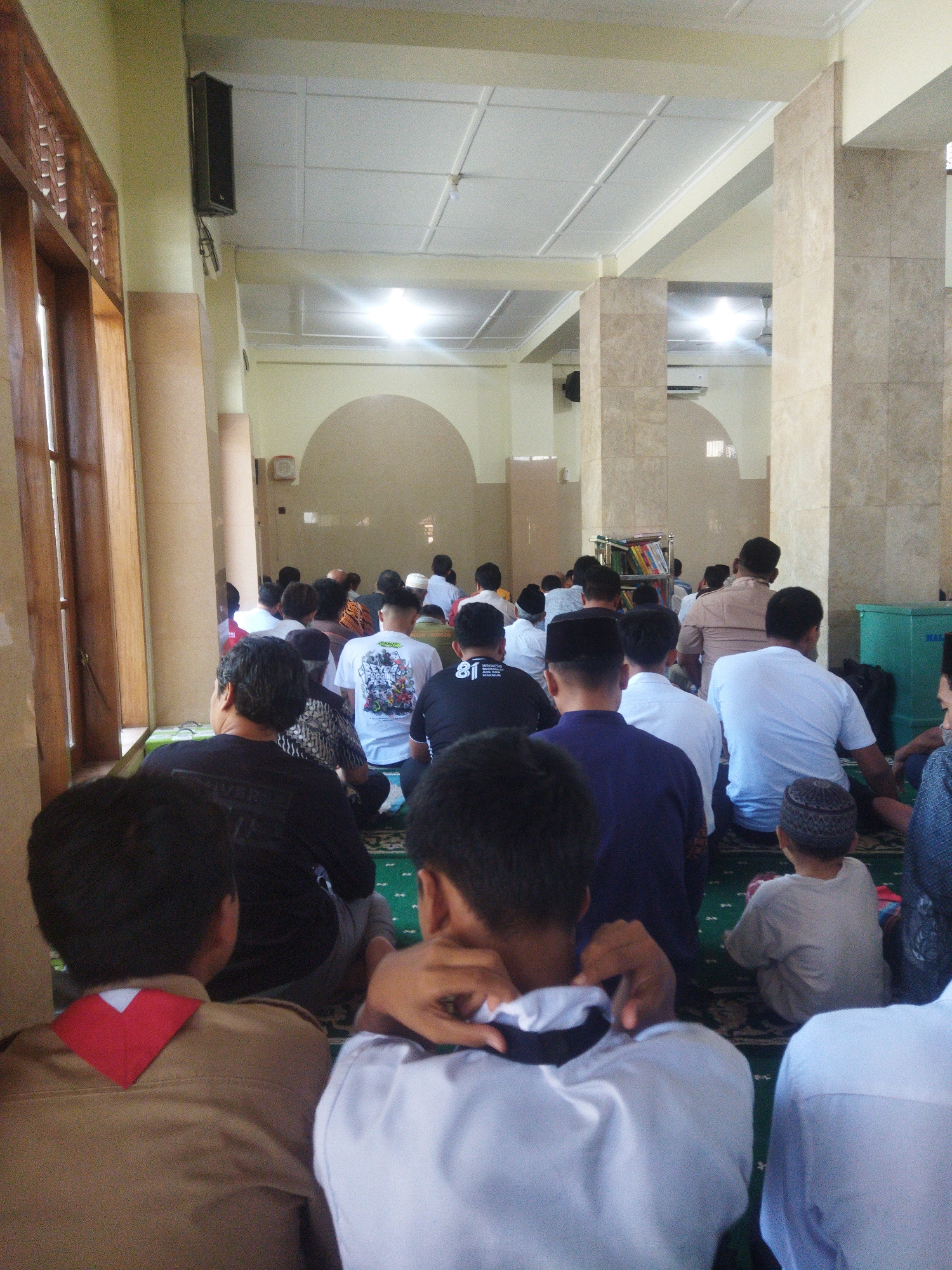 The image shows a view from behind a group of people sitting on a green carpeted floor, likely in a mosque. The individuals are facing away from the camera, towards an area where other people are also seated or in prayer. The room has a light yellow ceiling and walls, with large beige tiled columns providing architectural support. A wooden window with lattice work is visible on the left side of the frame. In the background, more people are seen, and there are also shelves with books, an air conditioning unit, and a green plastic bin. The lighting suggests it is daytime, with artificial lights also illuminating the space. The overall impression is of a religious gathering or prayer service.