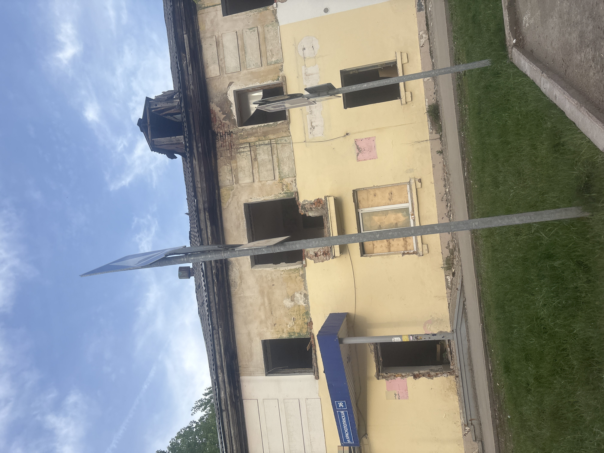 An eye-level shot captures the side of a dilapidated building in Perm, Russia. The building features a yellow exterior and is in a state of disrepair with multiple exposed window openings, some with brick visible. Several windows are boarded up. A blue street sign on a metal pole is attached to the building, which reads "ул. КРАСНОПОЛЯНСКАЯ 36." The sky is visible in the upper left, with white clouds. Grass and a concrete curb are visible to the right. The top of the building is damaged, revealing internal wooden structures.