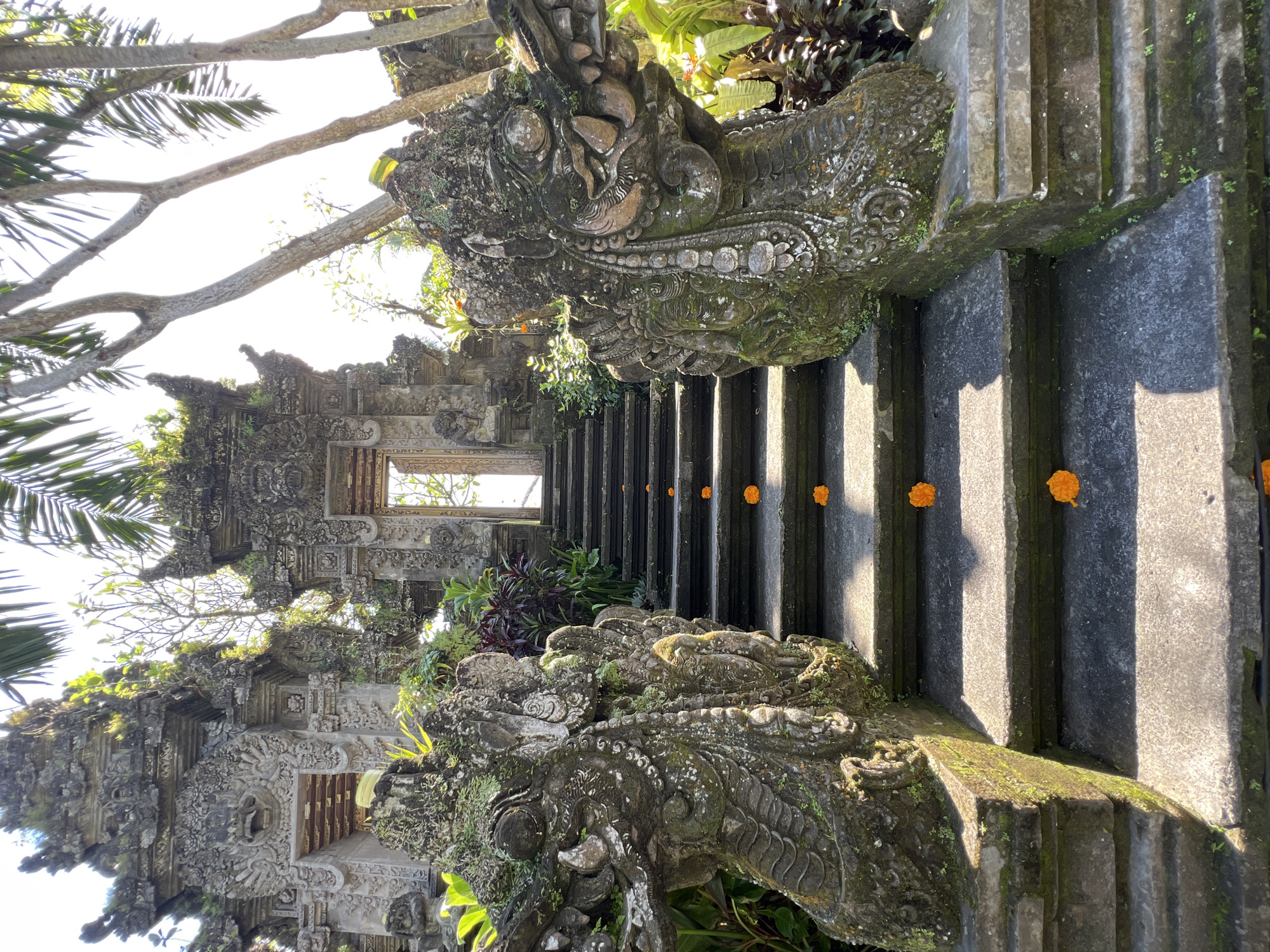 A stone staircase in Kedewatan, Indonesia, leads to a traditional stone building. Sculpted dragon-like figures, covered in moss and plants, flank the staircase. Orange flowers are placed on the steps. The architecture of the building is ornate, with detailed carvings. The scene is bathed in sunlight.