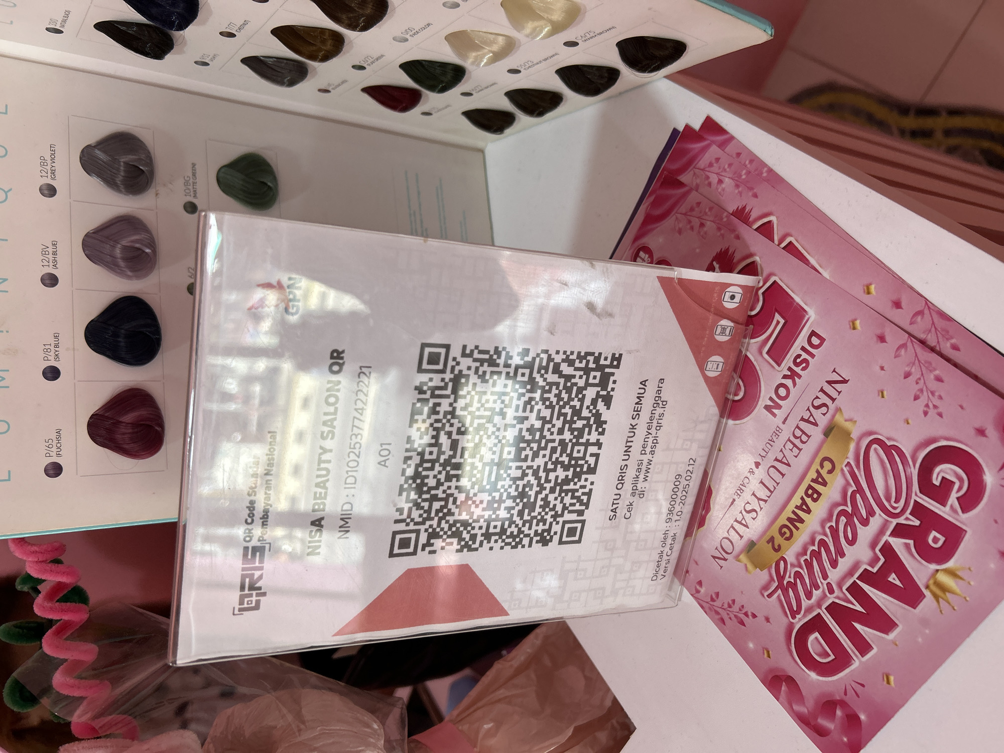 In the city of Depok, Indonesia, a close-up shot captures a display stand for a beauty salon. The stand prominently features a QR code for national payment, identified as "QRIS." Text on the stand reads "NISA BEAUTY SALON QR" and "NMID: ID1025377422221." Below the QR code, instructions state, "SATU QRIS UNTUK SEMUA" (One QRIS for All) and provide a website for checking the organizer's application: "www.aspi-qris.id." Additional details at the bottom indicate "Dicetak oleh: 93600009" (Printed by: 93600009) and "Versi Cetak: 1.0-2025.02.12" (Print Version: 1.0-2025.02.12).
Beneath the QR code display, several pink promotional flyers are visible. One flyer, partially obscured, displays the text "GRAND Opening" in large, bold font, with "CABANG 2" and "NISABEAUTYSALON" also legible. The flyers are adorned with decorative floral and leaf patterns.
In the background, to the right of the QR code stand, a display panel showcases various hair dye swatches. These swatches are labeled with names such as "P/65 (FUCHSIA)," "P/81 (SKY BLUE)," "12/BV (ASH BLUE)," and "12/BP (GREY VIOLET)," indicating different hair color options. The background also shows glimpses of decorative items, including pink pipe cleaners twisted into shapes. The overall setting appears to be an indoor commercial space, likely a salon or retail environment.