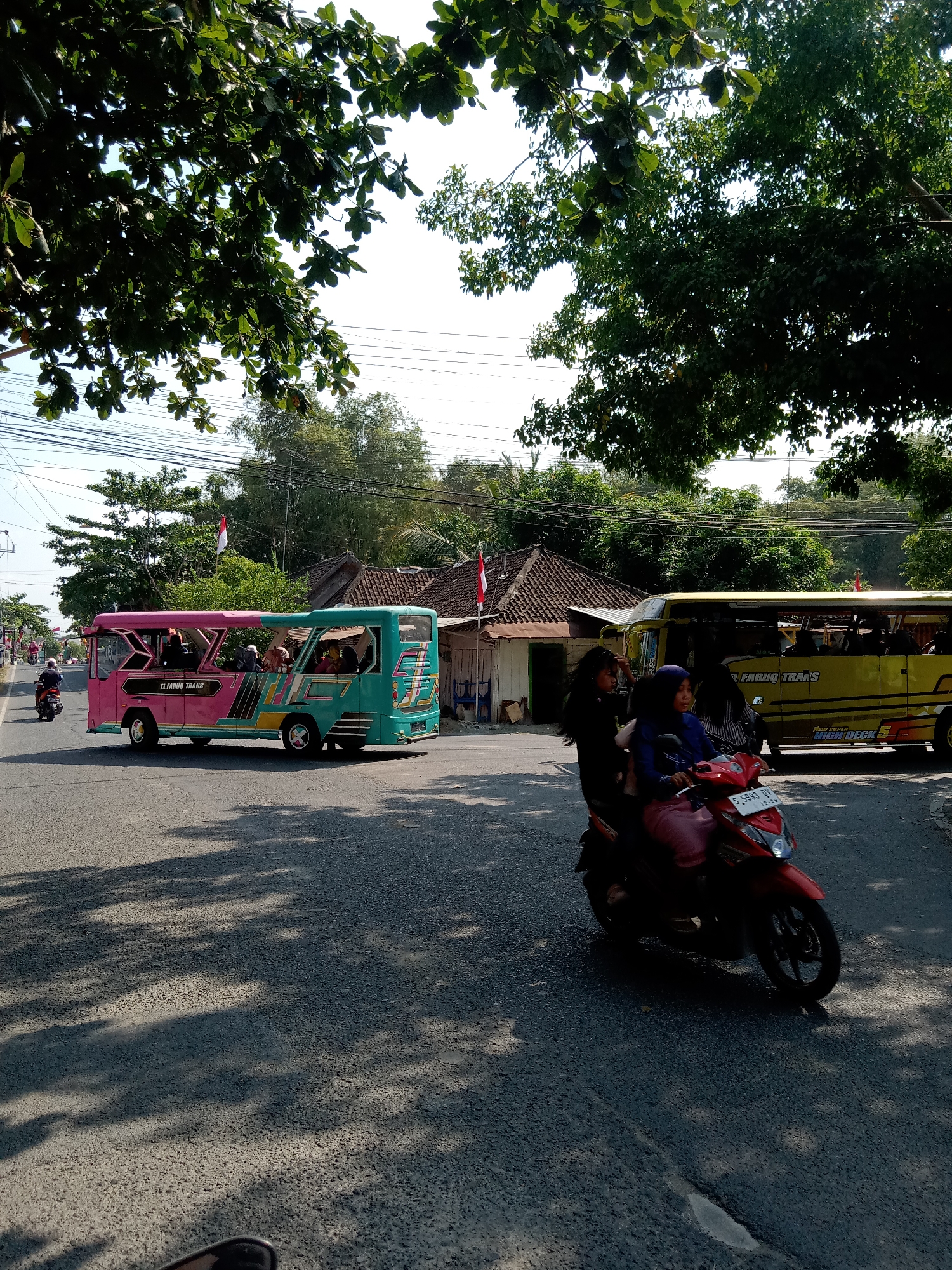 In Siwalan, Indonesia, on a sunny day, a dual-colored pink and teal bus, labeled "EL FARUQ TRANS," is seen at an intersection with a yellow bus. The pink and teal bus is transporting several passengers, including a person in the driver's seat. A red scooter with two passengers is approaching the intersection, with the rider wearing a dark blue hijab and a passenger in a pink headscarf. The environment is characterized by a paved road, shadows cast by trees, and a clear sky. In the background, houses with tiled roofs and trees line the roadside, with Indonesian flags visible on poles. The yellow bus also has passengers and displays the text "NEW SUPER HIGH DECK."
