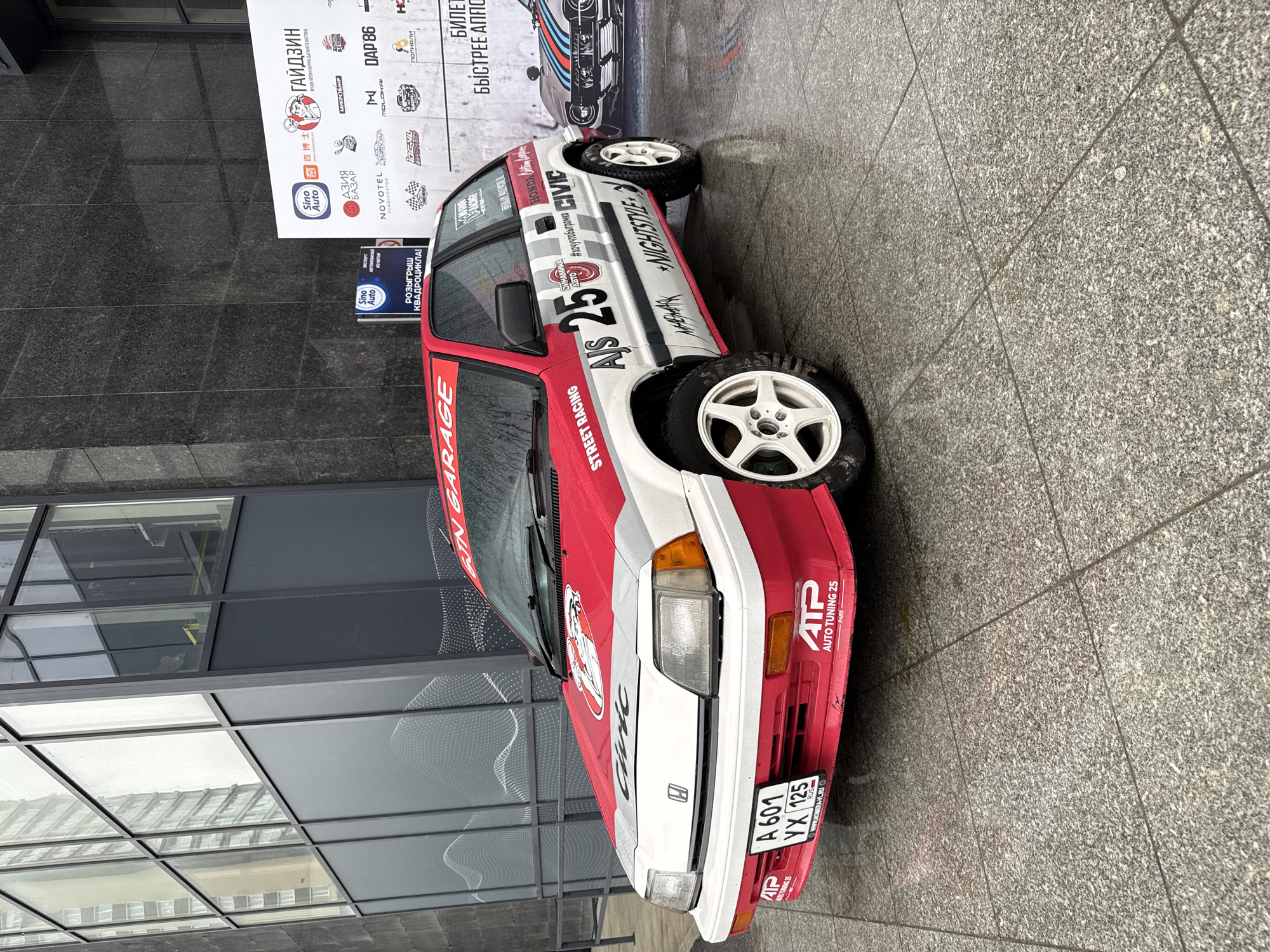 A red and white Honda Civic with racing decals is parked in front of a building with glass windows and a gray stone facade. The car's license plate reads "A 601 YX 125" and indicates that the vehicle is from Russia. A banner in the background advertises "GIN GARAGE," various sponsors, and a contest to win a quad bike, referencing Sino Auto and the Novotel Vladivostok. The banner also mentions "GAIDZIN," the Museum of Automative Culture of the Far East. The surface is wet, suggesting it has recently rained.