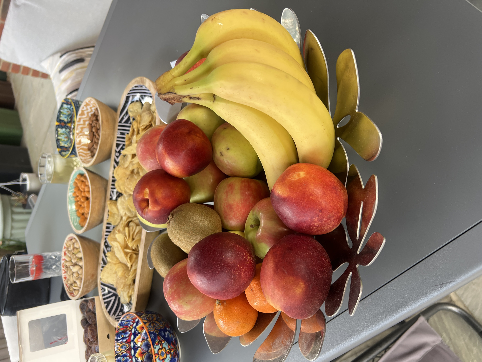 This is a medium shot, eye-level, close-up photograph of a fruit bowl on a grey outdoor table. The fruit bowl, which appears to be made of brushed metal with a leaf-like design, contains a variety of fruits including bananas, apples, nectarines, kiwis, and tangerines. In the background, there are several bowls filled with snacks such as pistachios, crisps, and nuts. There are also drinks in glasses and a box of dates visible. The setting suggests an outdoor gathering or party. The image does not contain any people and no specific landmarks identifiable to Guildford, United Kingdom are present.