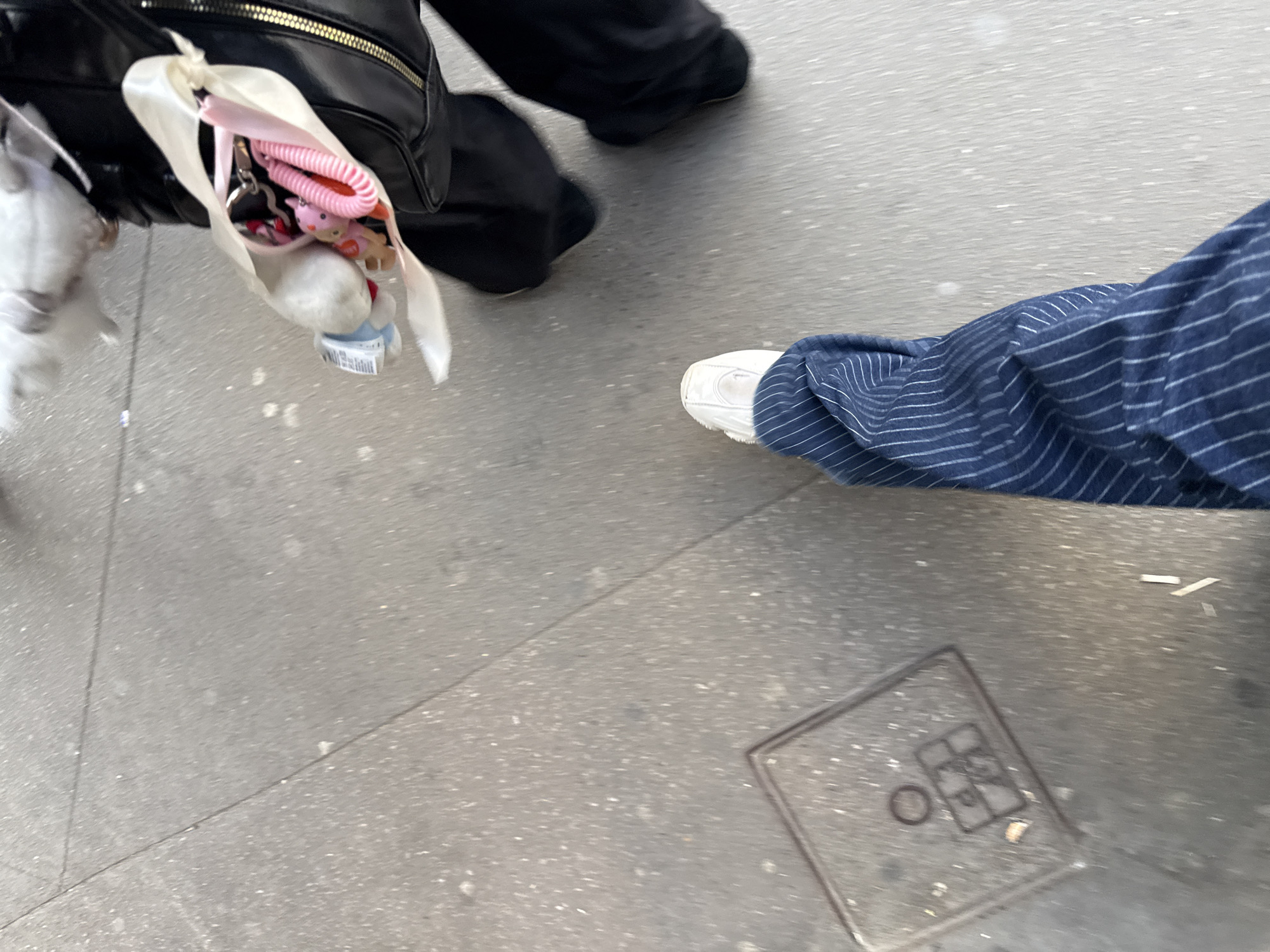 This low-angle photograph captures a street-level view of two individuals walking or standing on a light gray urban pavement, likely in Paris, France. The slight motion blur suggests movement.
On the left side of the frame, a person is visible wearing dark, likely black, wide-leg pants or a skirt. A black bag with a prominent gold zipper is slung over their shoulder. Attached to this bag is a small, soft white pouch, which contains several items: a pink coiled accessory (possibly a hair tie or keyring), a small pink plush toy, and a distinct white plush character, which appears to be Hello Kitty due to a visible red bow and general shape. A small blue tag with unreadable text is visible beneath the white plushie.
On the right, the lower leg and foot of another person are visible. They are wearing blue pants with thin white vertical stripes, bunched slightly at the ankle, and a pair of white athletic sneakers with textured soles.
The pavement itself is a speckled gray, showing some cracks and lines. In the lower right corner of the image, a square metal utility grate is embedded in the ground. It features etched symbols, including a circle and a square with an internal cross pattern, but no legible text. The scene appears to be taking place during the daytime, given the general lighting, though no direct light source or strong shadows are present.