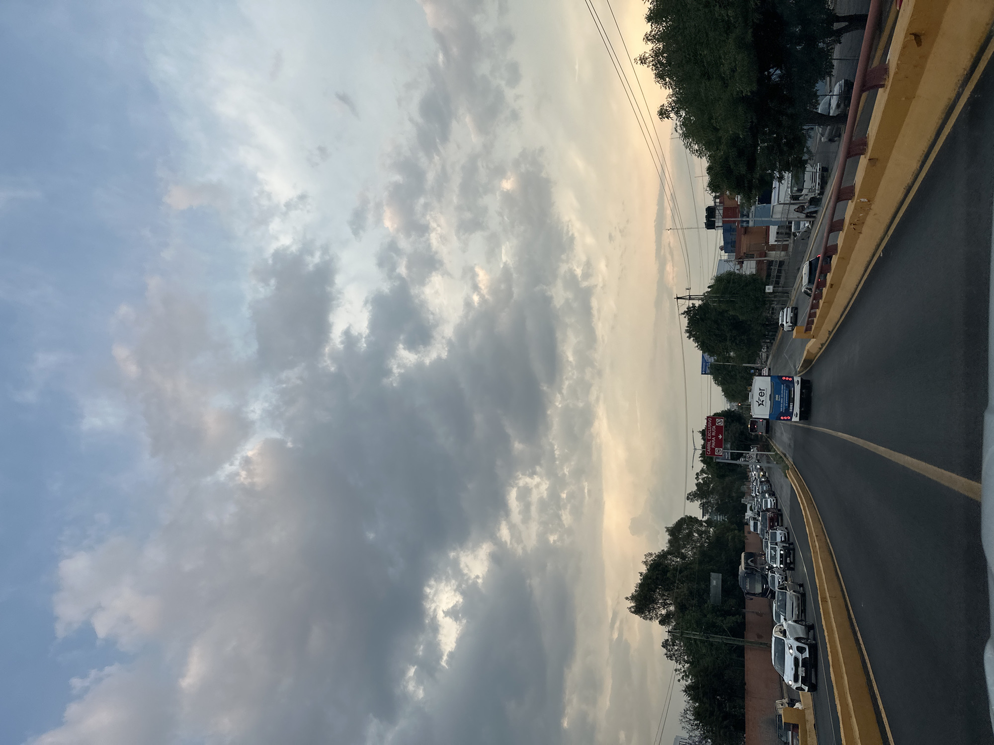 This vertically oriented image captures an elevated road or overpass, likely a major thoroughfare, in Puebla, Mexico, during twilight hours, possibly late afternoon or dusk. The sky dominates the upper portion, displaying a mix of light blue and heavy grey clouds, with brighter patches indicating soft, warm light from the sun either setting or rising on the horizon.
The scene depicts a busy traffic situation. On the right side of the asphalt road, numerous vehicles, including sedans, SUVs, and a prominent white Ford Focus, are caught in what appears to be a traffic jam or very slow-moving traffic. A white and blue bus, with "Xpr" or "EXPRES" (partially visible) printed on its rear, is at the head of the visible traffic queue. The road features clear yellow painted lines and a distinct yellow concrete barrier separating lanes or the shoulder.
On the left side of the frame, behind the yellow barrier, a line of green trees partially obscures buildings and structures further in the background. Faint utility poles and power lines are visible against the cloudy sky. A red-on-white road sign is noticeable among the trees, with partially legible text reading "CARRIL EXCLUSIVO PARA..." (Exclusive Lane For...). The overall atmosphere is that of a bustling urban environment at the transition between day and night, under an overcast sky.