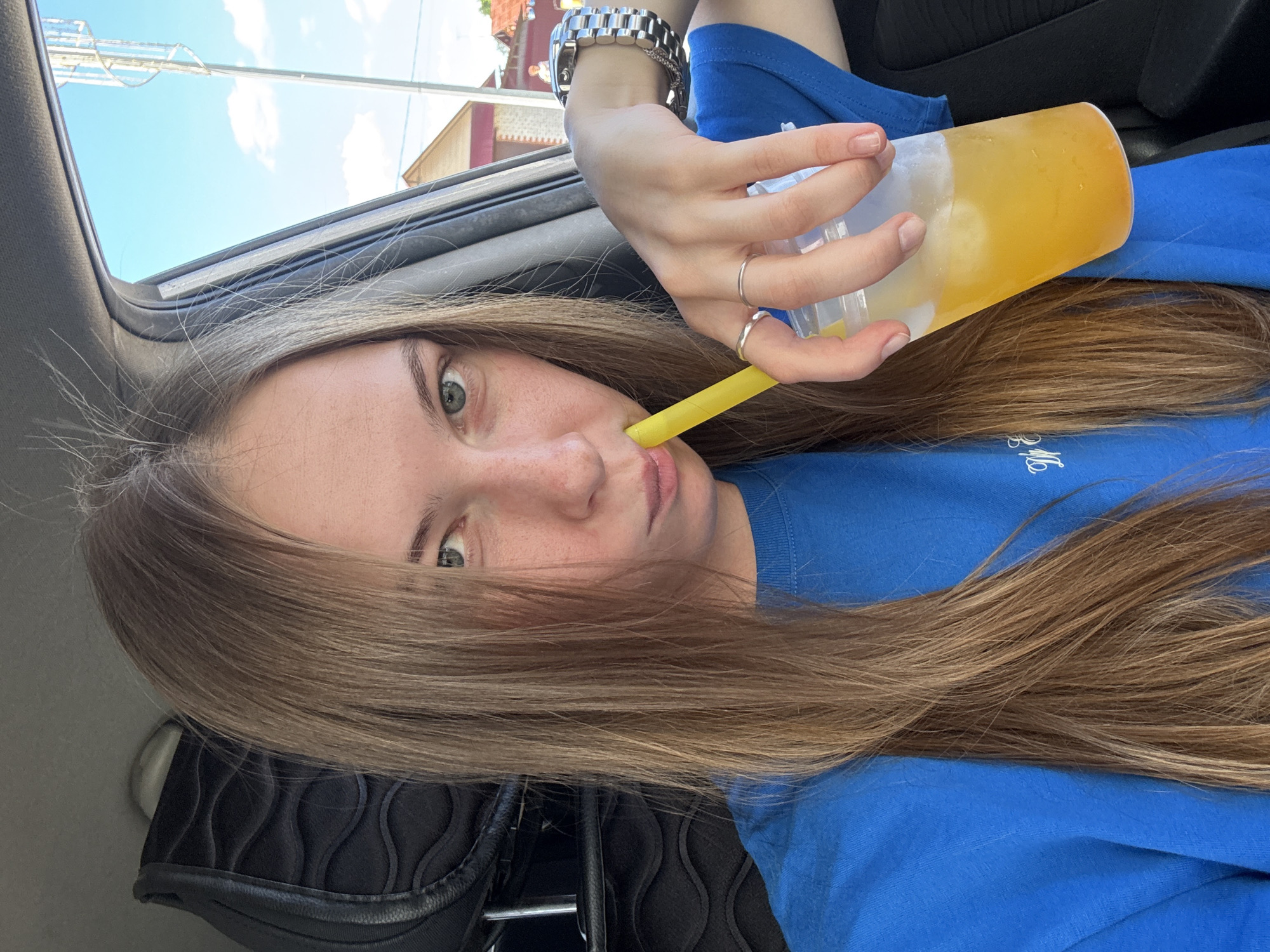 This media file features a young woman inside a vehicle. She has long, light brown hair that is spread across her shoulders and lap. Her head is tilted to the side as she drinks from a clear plastic cup filled with a yellow beverage and ice, using a yellow straw. She is wearing a bright blue t-shirt and several rings on her fingers, along with a silver watch on her left wrist. The interior of the car is partially visible, including the headrest of a black car seat and a portion of the car's door panel. Sunlight streams in from an open sunroof, indicating it is daytime. The sky, visible through the sunroof, is blue with scattered white clouds. In the background, out of focus, are buildings and what appears to be a light pole with a structure on top, suggesting an urban or suburban environment. The overall mood appears casual and relaxed, possibly during a car journey on a sunny day. There is some faint white text visible on the blue t-shirt, but it is not clearly legible. The context suggests this photo was taken in Perm, Russia.