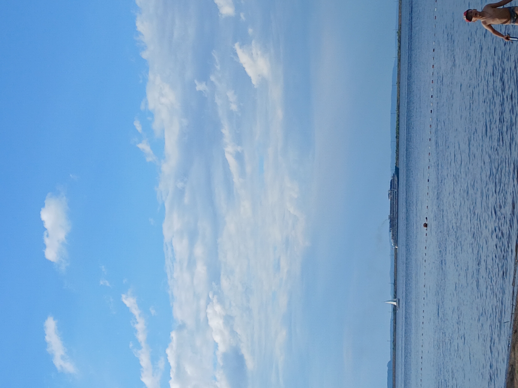 This media file depicts a scenic view of a body of water under a bright blue sky filled with scattered white clouds. In the foreground, on the right side of the frame, a young person wearing a red cap and swim trunks is visible from the waist up, looking towards the water. They appear to be standing at the edge of the water. The water itself is calm and reflects the sky, with subtle ripples. A line of buoys or markers stretches across the water, indicating a designated swimming or boating area.
In the distance, a white sailboat can be seen navigating the water. Further back, a large ship or ferry is visible on the horizon, along with a distant shoreline featuring some buildings and what appears to be a landscape of hills or mountains. The sky suggests it is daytime, likely a clear and pleasant day. The overall atmosphere is peaceful and suggests a recreational setting, possibly a beach or a lake. The image is oriented vertically, with the sky occupying the left half and the water and distant scenery on the right. There is no visible text in the image.