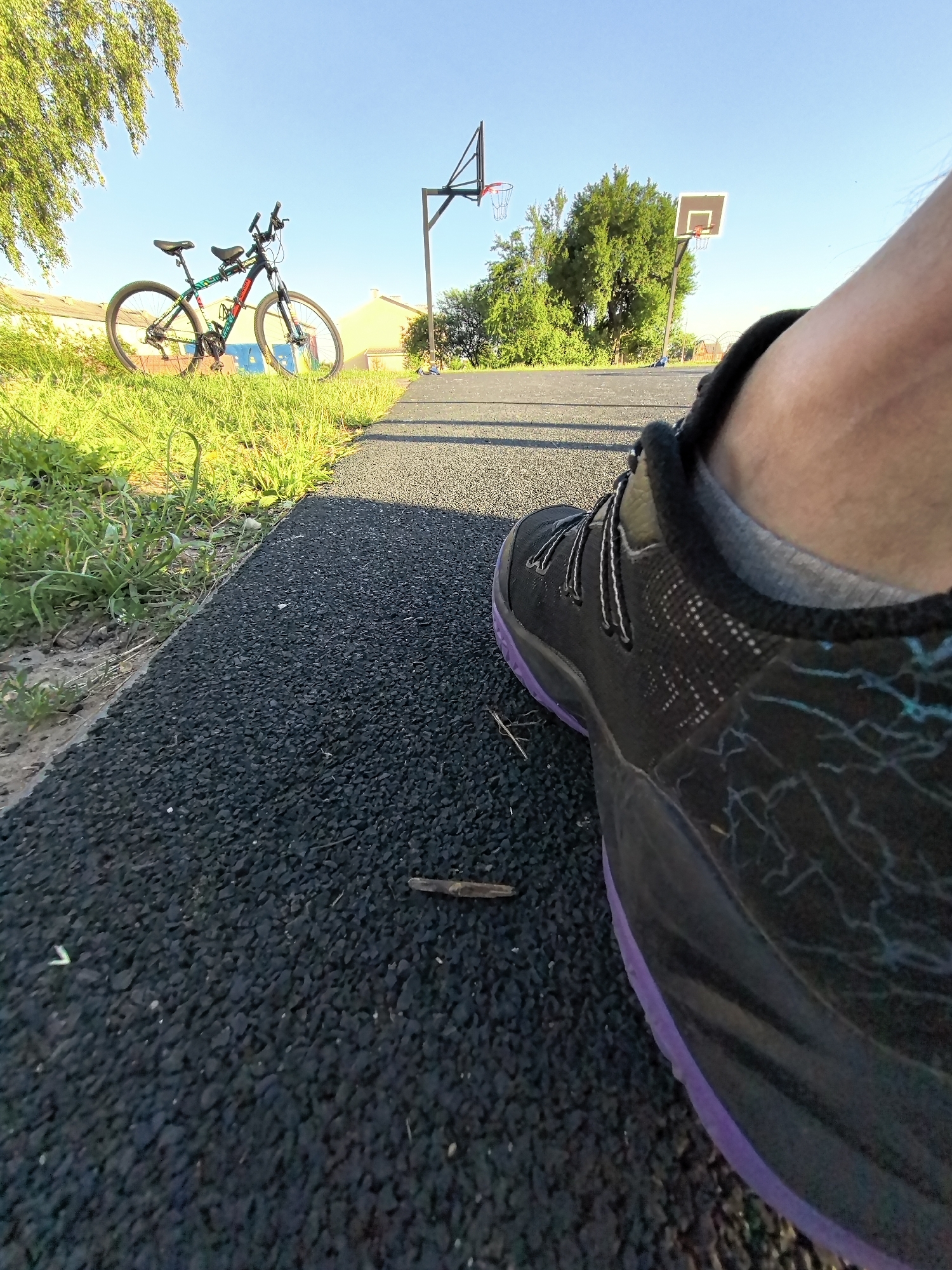 The image shows a low-angle, close-up view of a person's foot wearing a black athletic shoe with purple accents, standing on a dark, textured surface. In the background, a bicycle with colorful accents is parked on a grassy area. Beyond the bicycle, there are two basketball hoops, one with a red rim and net, and another white backboard with a red rim and net. Residential buildings with yellow and beige exteriors are visible behind the basketball hoops, along with several green trees. The sky is clear and blue. The scene appears to be an outdoor recreational area, possibly a park or a schoolyard, on a sunny day.