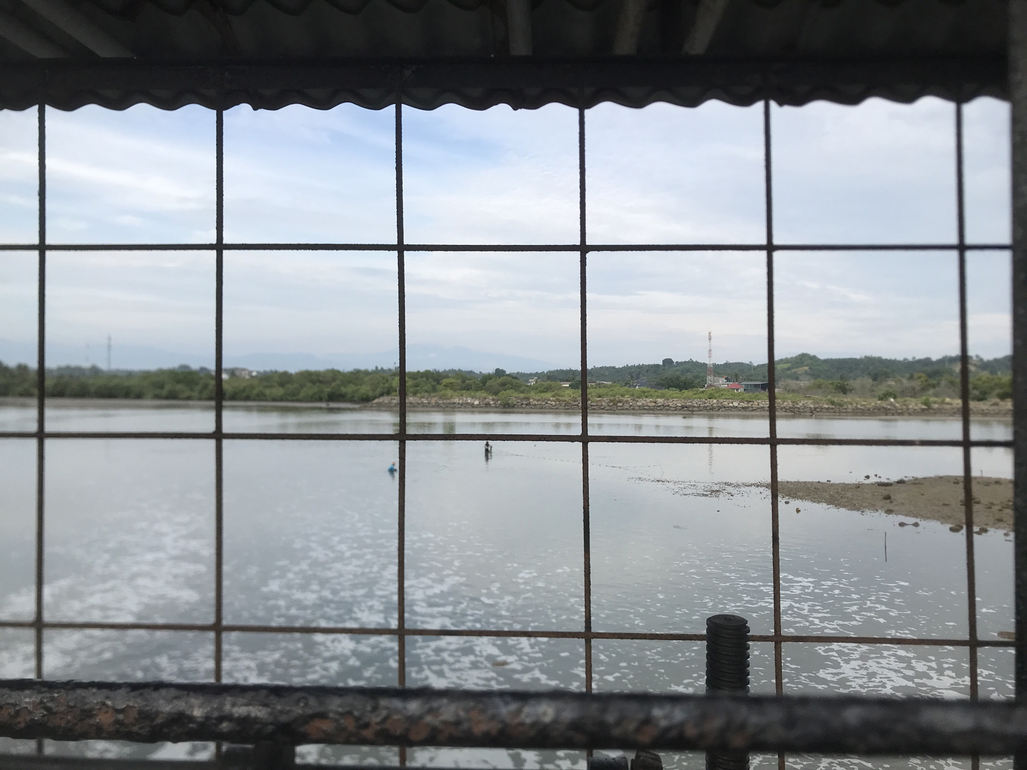 The image shows a view of a body of water, possibly a river or bay, seen through a rusty metal grate. The water has a slightly choppy surface with white foam in some areas. In the distance, the opposite bank is lined with trees and some structures, including what appears to be a telecommunications tower. There are two small figures in the water, one is a silhouette of a person standing in the water, possibly fishing or wading, and another smaller blue object which could be a buoy or another person. The sky is overcast with scattered clouds. The foreground is dominated by the metal grate, which is weathered and shows signs of rust. The overall scene suggests an outdoor, possibly industrial or waterfront location. While the image does not explicitly show a landmark identifiable as being specifically within Lhokseumawe, the description of the scene is consistent with coastal or riverine environments found in such Indonesian cities.