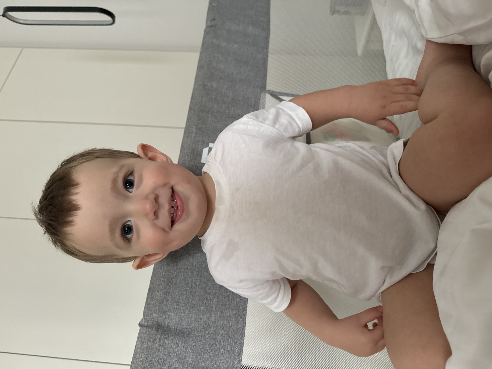 A young child lies in a crib. The child is wearing a white T-shirt and appears to be wearing a diaper. The crib has a gray side rail. The background includes a white wall and what appears to be bedding. No information indicates this image was taken in Odintsovo, Russia.