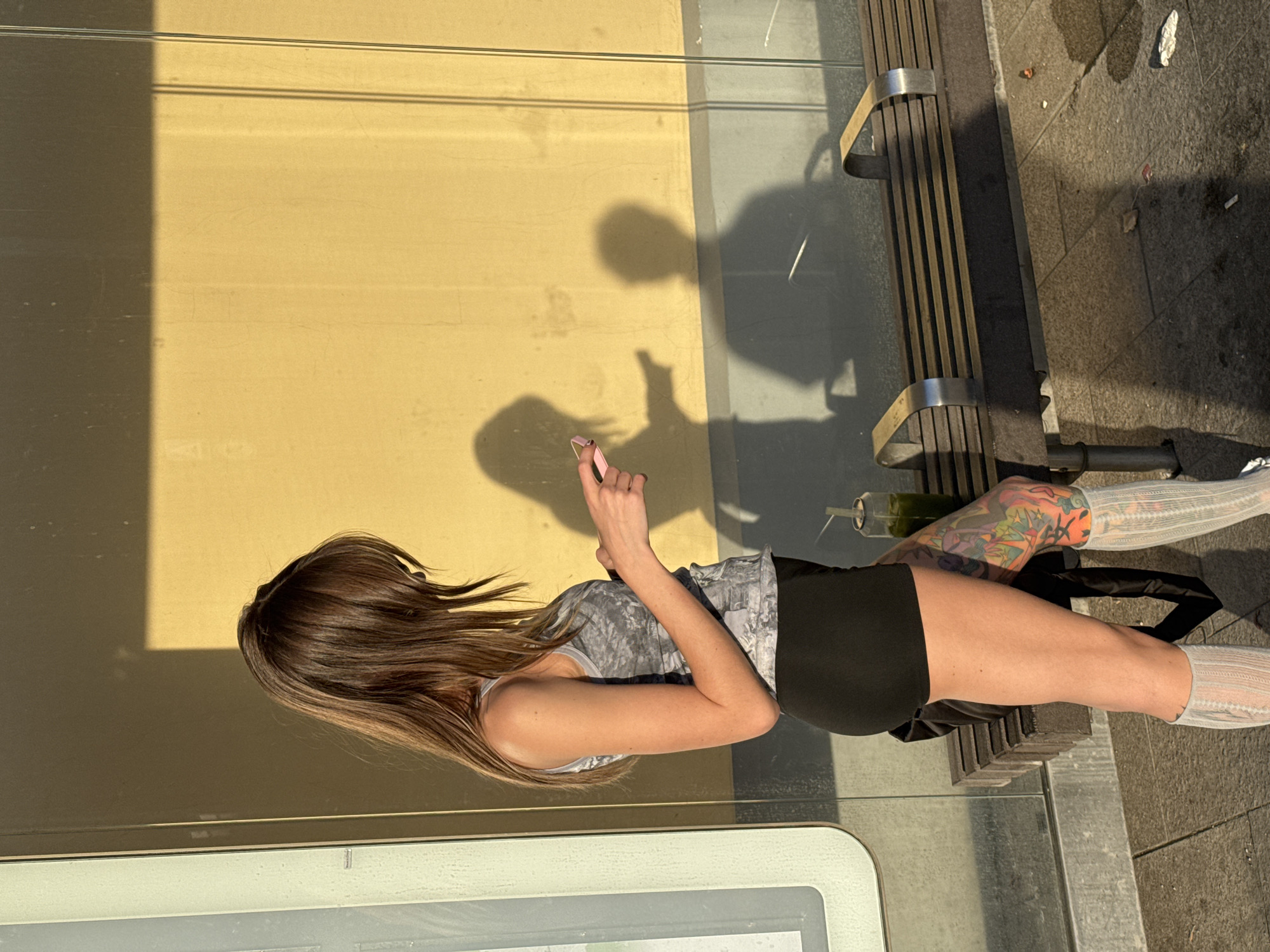 A woman stands in profile, looking down at a pink smartphone she holds in her hands. She is wearing a grey, patterned tank top, black shorts, and knee-high grey socks. Her left leg has elaborate colorful tattoos. She is standing on a sidewalk, next to a bus stop bench with a glass enclosure behind it. Sunlight casts long shadows on the yellow wall of the bus stop. A green drink in a clear plastic cup sits on the bench. The ground is paved with square tiles, and there are scattered pieces of litter on the ground. The scene is outdoors, and appears to be in an urban environment. The bus stop appears to be located in Moscow, Russia.