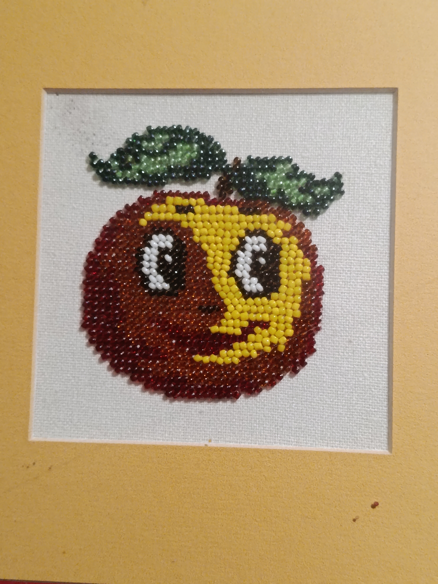 The image is a framed, beaded artwork of a stylized apple with a face. The apple is predominantly red and yellow, with two green leaves attached to the top. The face features two large white eyes with black pupils and a smiling mouth formed by red beads. The artwork is mounted on a white textured mat and framed by a yellow border. The artwork is not located in Chișinău, Moldova.