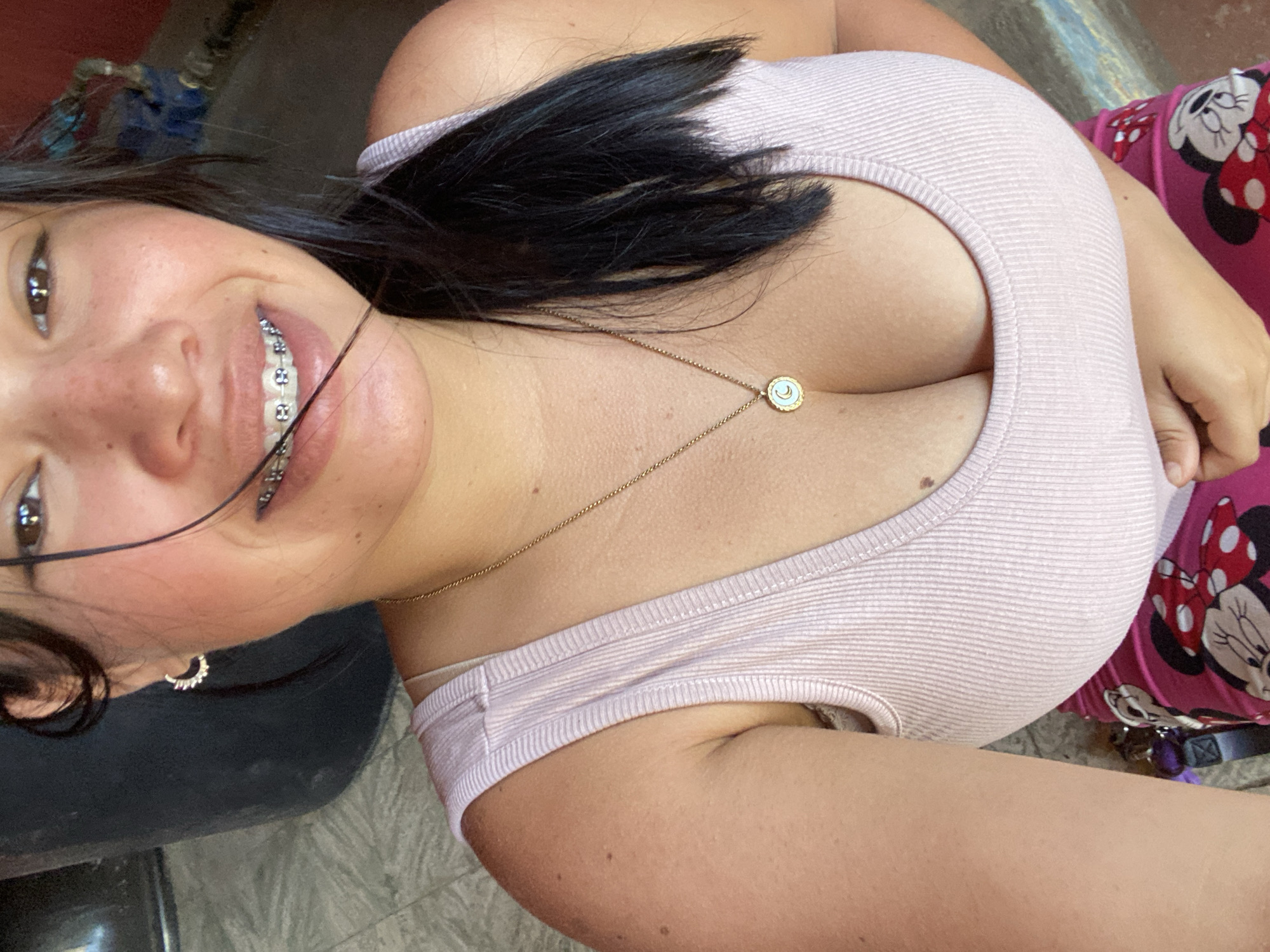 A selfie shows a young woman with dark hair, braces, and a gold necklace wearing a pink tank top and pink Minnie Mouse pajamas. The setting appears to be indoors, possibly a private residence. There's a dark object to her left and a patterned surface underneath. There is no evidence to indicate the location is Juan Griego, Venezuela.