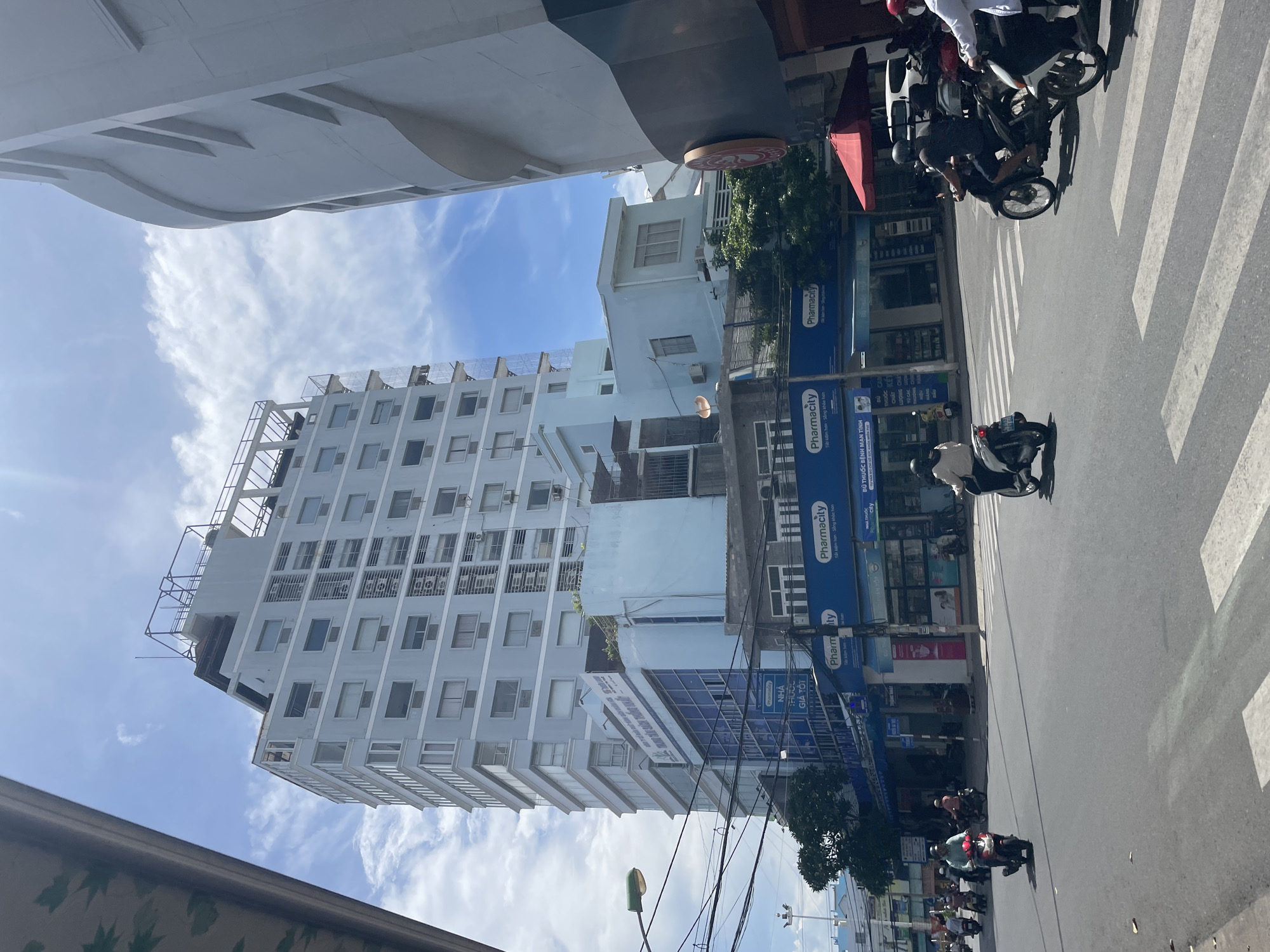 The image captures a daytime street scene in Nha Trang, Vietnam, dominated by a tall, light blue, multi-story building. The building features numerous regularly spaced windows and visible air conditioning units. To the right of this structure is a partially visible, larger, curved, white building. The street in the foreground is paved, with white pedestrian crossing lines visible. Several people are riding motorbikes. One individual in a light green shirt is on a red motorbike in the left lane, while two individuals are on dark-colored motorbikes in the right lane, approaching the pedestrian crossing. A person on a white motorbike is in the left lane, further back, also heading towards the crossing. The buildings lining the street have prominent signage, including "Pharmacity" in blue and white, indicating pharmacies. There are streetlights and overhead wires present. The sky is blue with scattered white clouds, and sunlight is casting shadows, suggesting a sunny day.
