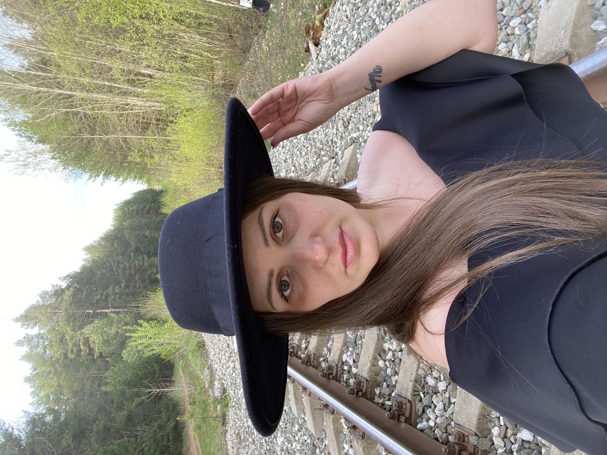 A woman wearing a black hat and a dark top is positioned on a railroad track. She rests her hand on the hat. The background includes trees and foliage. The location is Chaikovsky, Russia.