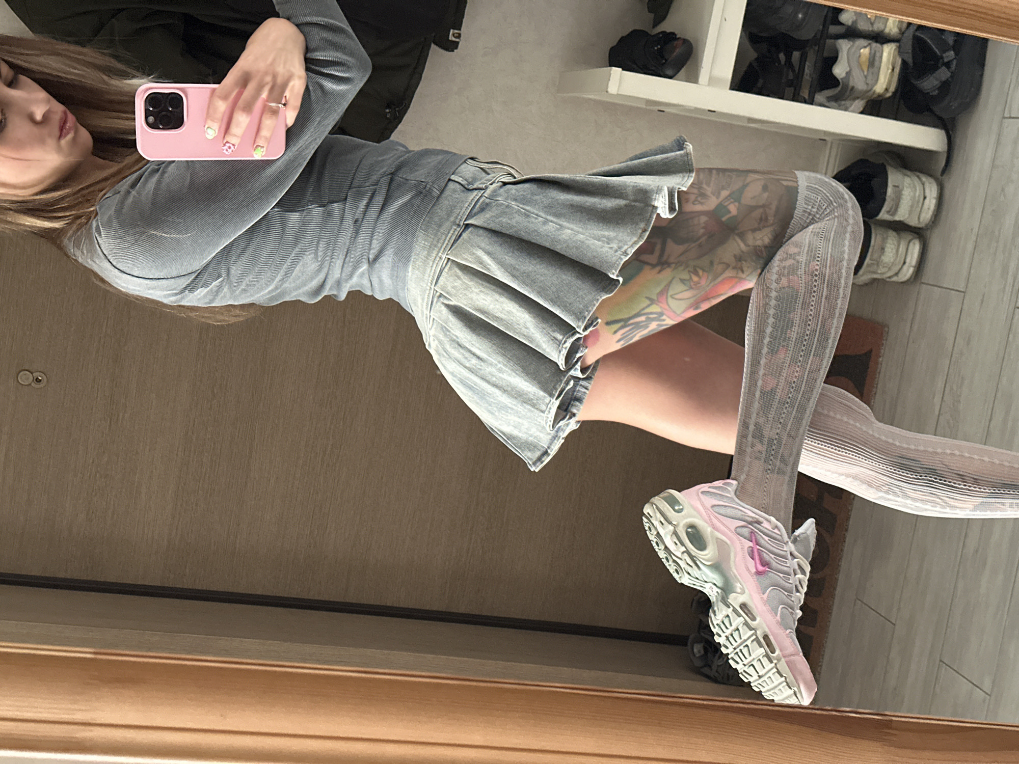 A young woman is taking a mirror selfie in what appears to be a home setting, possibly a hallway or dressing area given the wooden door and flooring. She is wearing a light blue, ribbed long-sleeved shirt and a pleated denim skirt. Her left arm is raised, holding a pink smartphone, which is angled to capture her reflection. She has long, blonde hair and visible tattoos on her right leg. She is also wearing white, patterned thigh-high socks and light pink and white athletic shoes. The background shows a shelf with various shoes and a dark jacket hanging. The lighting suggests it is daytime, possibly indoors with artificial light. There is no visible text in the image. The overall mood is casual and stylish.