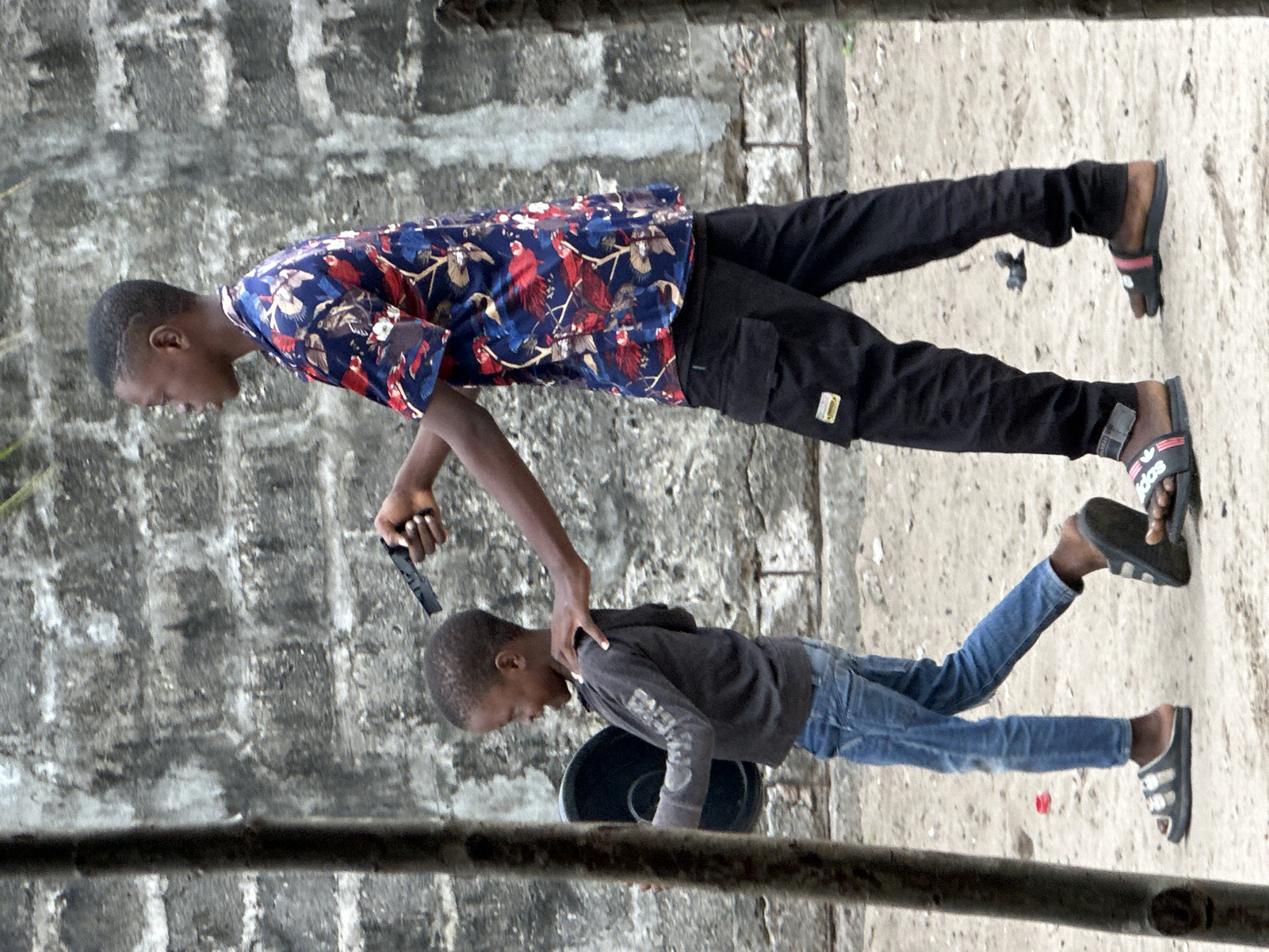 In Badagry, Nigeria, a young Black male, appearing to be a teenager, stands to the right of the frame. He is wearing a colorful short-sleeved shirt with a floral and bird pattern in shades of blue, red, and white. His lower half is clad in black cargo pants with multiple pockets, and he wears black slide sandals with pink accents. In his right hand, he holds a black handgun, pointed forward and slightly downward, with his index finger near the trigger. His left arm is extended, resting on the shoulder of a younger Black male to his left.
The younger Black male, who appears to be a child, is facing left and walking away from the viewer. He has short, dark hair and is wearing a dark gray hooded sweatshirt with some light-colored text or graphics on the sleeves. He is also wearing blue jeans and dark gray slide sandals. In his left hand, he carries a circular, dark-colored object, possibly a bowl or plate, held against his body.
They are walking on a sandy or dirt ground. Behind them is a textured, grey concrete wall that appears weathered, with visible blocks and some staining. To the left of the frame is a thick, dark vertical post, possibly a tree trunk or wooden pole, which partially obscures the younger boy. Another similar post is visible on the far right edge of the frame. The overall lighting suggests an overcast or diffused daylight condition.