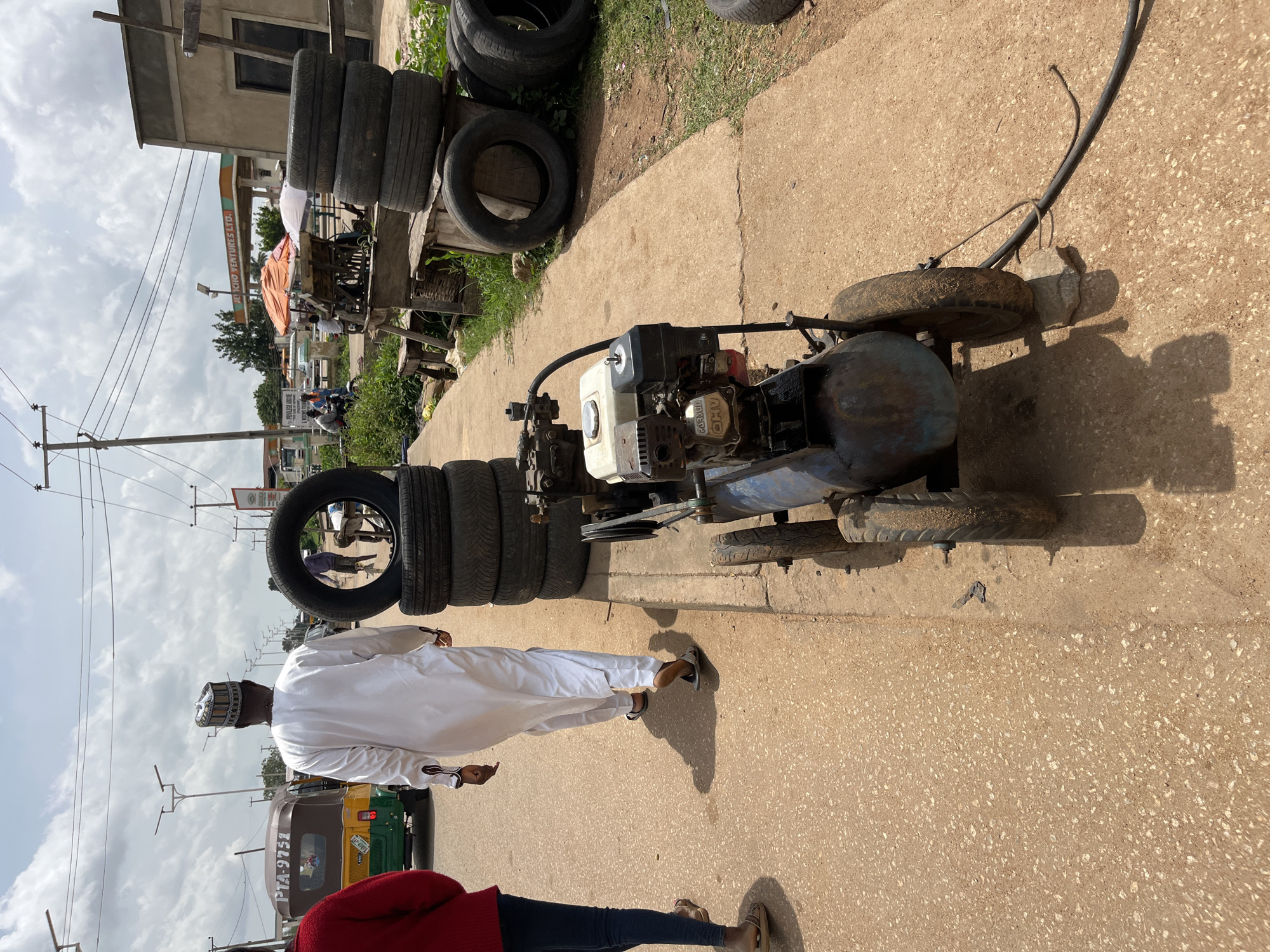 In Bukuru, Nigeria, an outdoor scene depicts a man in traditional white attire and a patterned cap walking away from the viewer, his shadow cast on a dusty, concrete path. To his left, a red-clad woman and a person in jeans are also walking on the path. A yellow and green tricycle is parked nearby. Stacks of used tires are visible on the right side of the path, some piled high and others lying on the ground. A blue air compressor with a small engine, possibly a "Guanli" model, is positioned on the path, its wheels resting on small stones and a cable extending to the right. The background shows a gas station with an orange canopy, a building with a sign reading "JET CHO VENTURES LTD," and utility poles with overhead wires under a cloudy sky. The environment appears to be a roadside commercial area.