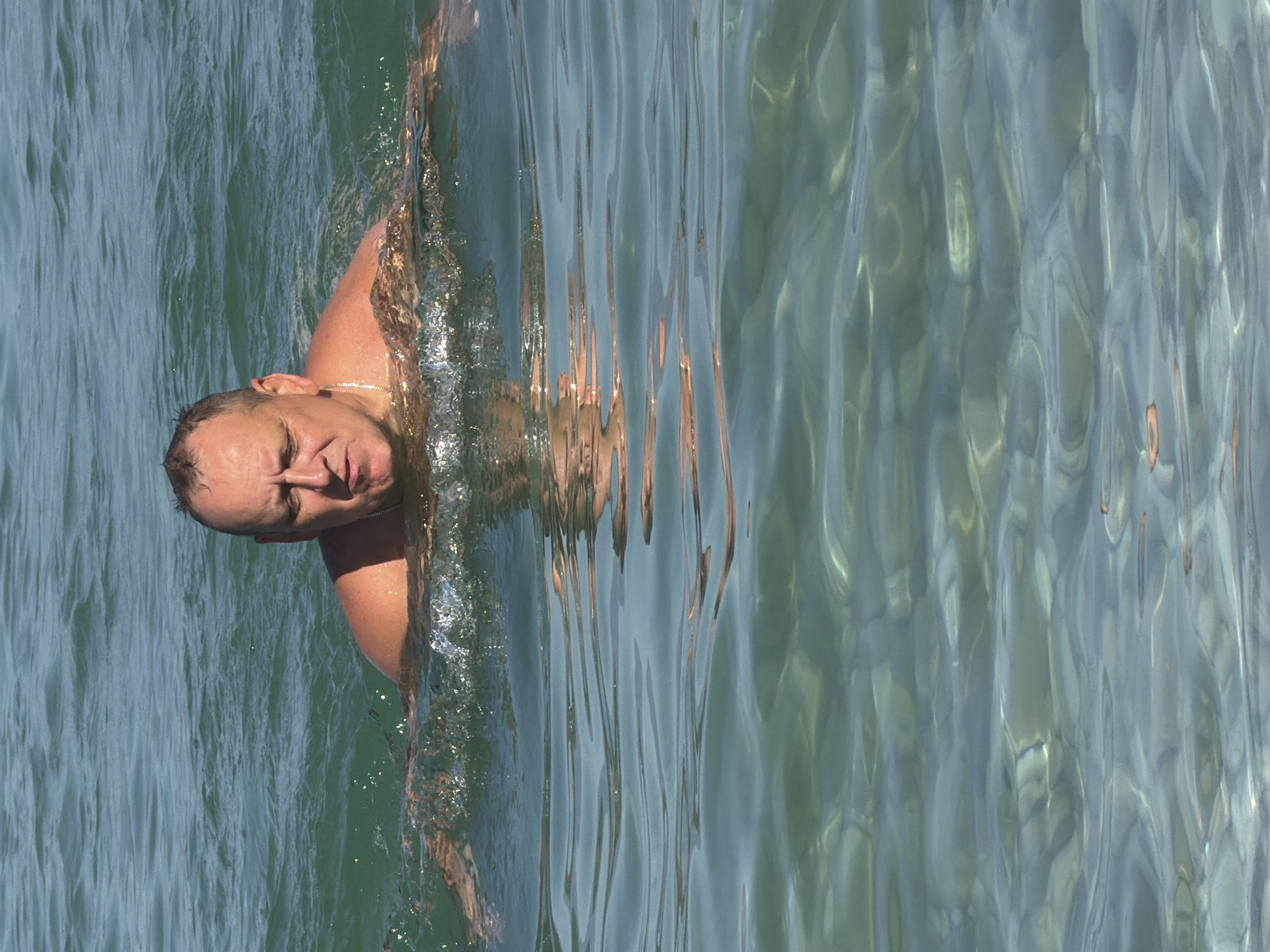 A middle-aged Caucasian man is swimming in the clear, turquoise waters of the Adriatic Sea, at a beach in Općina Baška, Croatia. The man is facing forward, with his head and shoulders above the water. He has short, greying hair, a fair complexion, and is wearing a thin gold chain around his neck. The water is calm, with gentle ripples reflecting the sunlight. The seabed is visible through the clear water, with scattered pebbles and sand. The overall scene is peaceful and serene, conveying a sense of relaxation and enjoyment of the natural environment.