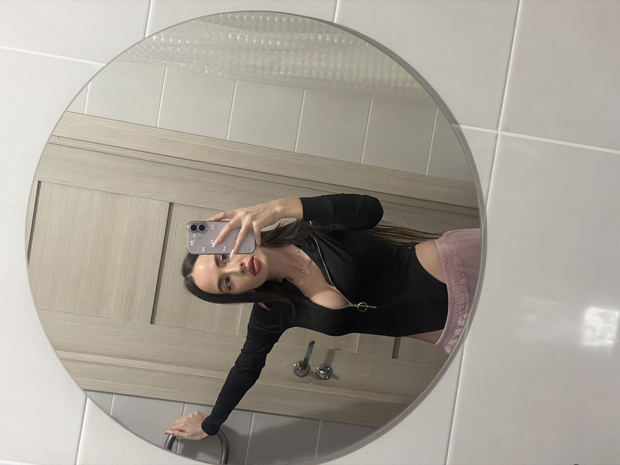 A woman is taking a selfie in a round mirror in what appears to be a bathroom. The wall behind the mirror is tiled in white with a light wood vanity. The woman is wearing a black long-sleeved top with a zipper and pink shorts. She is holding her phone up to take the picture, and her expression is neutral. The image does not contain identifiable features or indicators that would provide the location of Saint Petersburg, Russia.