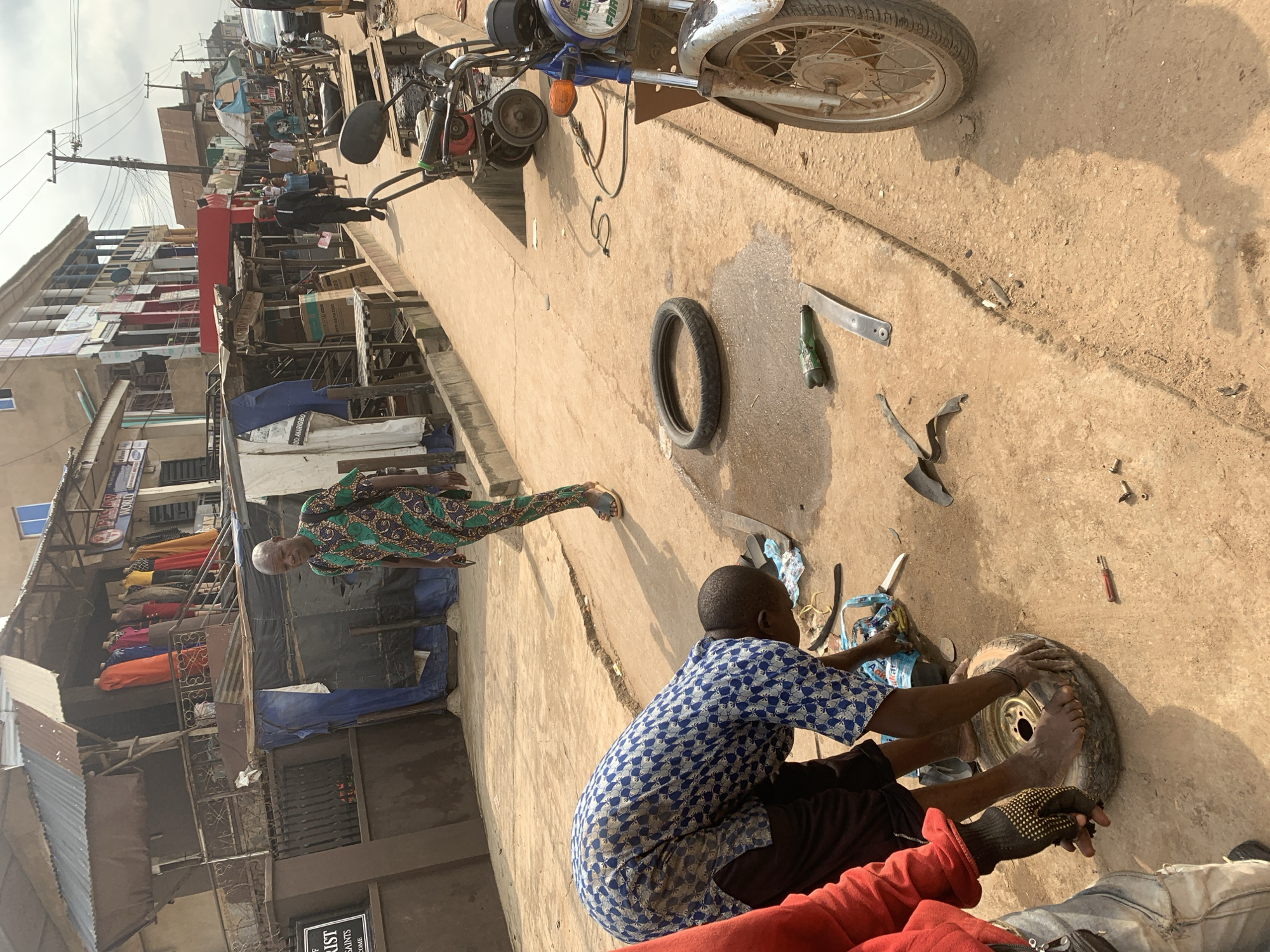 The image captures a street scene in Shagamu, Nigeria, where a man is working on a tire. He is bent over, with one foot placed on a tire, and his hands are engaged with it. Another person in a red shirt and dark gloves stands nearby, also seemingly involved in the task. Tools and tire parts are scattered on the dusty ground around them. In the background, a man in a colorful patterned outfit walks by, and various shops and stalls line the street, with motorcycles and other vehicles parked along the roadside. The environment suggests a bustling marketplace or commercial area.