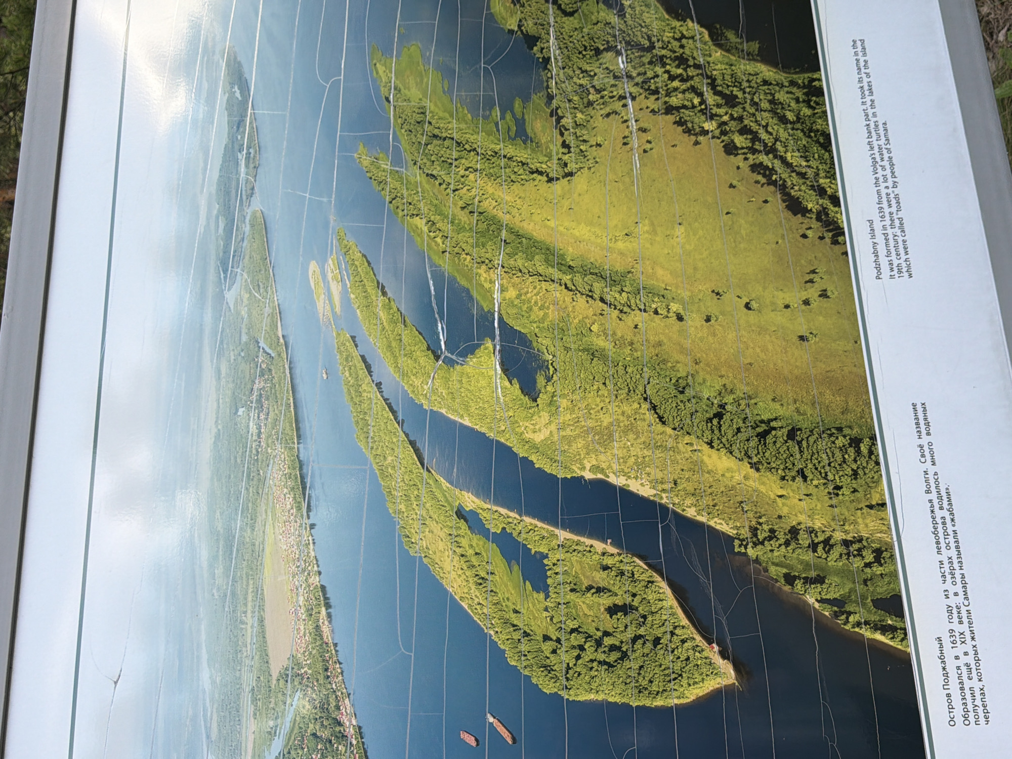 The image shows a framed informational plaque with a photograph of a wide, blue river with several long, green islands. The islands are covered in dense trees and vegetation, and the river has multiple channels. In the distance, a city or town can be seen on the left bank of the river, with scattered buildings and trees. Two cargo ships are visible on the river. The plaque has text in both Russian and English, identifying the scene as "Podzhabny Island" and stating that it was formed in 1639 from the Volga's left bank. The English text also mentions that the island's name comes from the abundance of water turtles found there, which were called "toads" by the people of Samara. The location is implied to be near Volzhskiy, Russia, given the context of the Volga River and the proximity to Samara. The photograph itself is of an aerial perspective, showcasing the geography of the river and its islands.