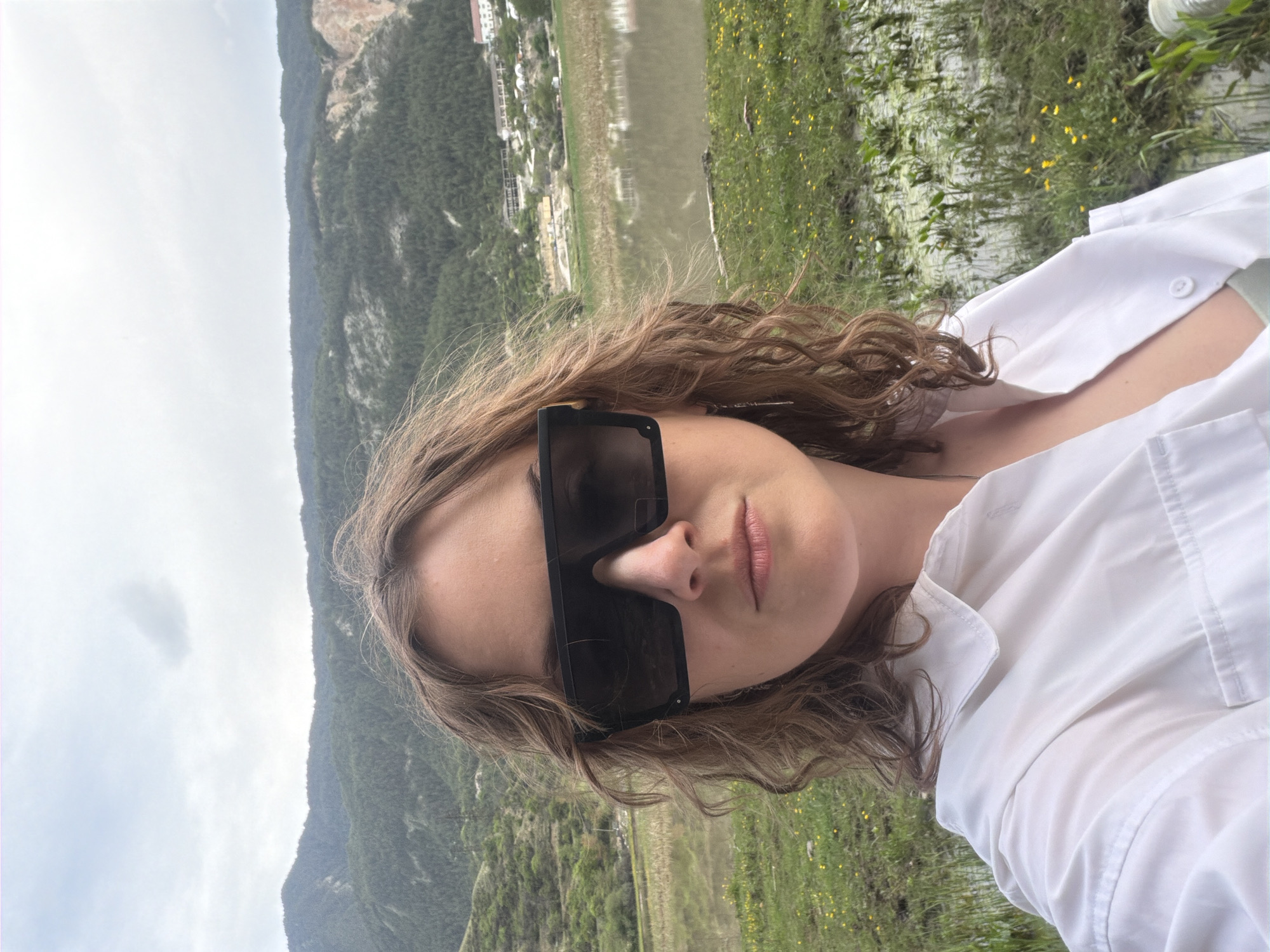 The image shows a woman wearing large black sunglasses and a white shirt, set against a scenic background. The background includes a mountain, a body of water, and some greenery with yellow flowers. A small town or village is visible in the distance near the water. The location is not identifiable from the given information.