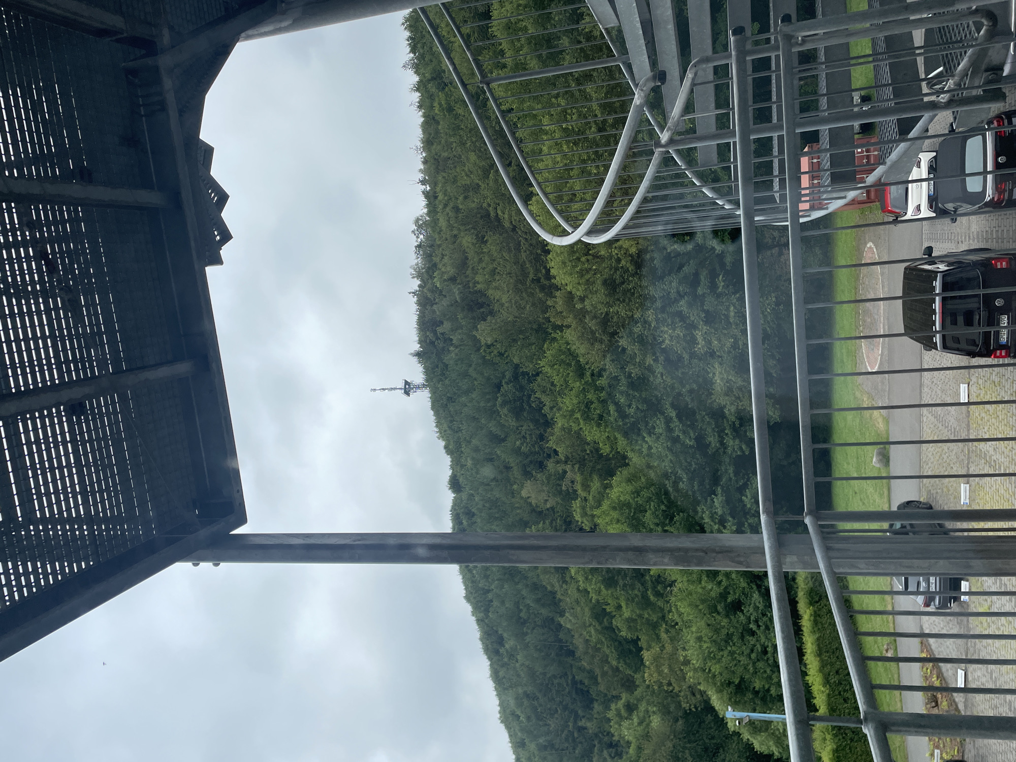 The image shows a view from a metal staircase structure, looking out over a heavily wooded hill with a radio tower on its crest. The sky is overcast. In the foreground, a grey spiral staircase is visible, along with a section of a metal walkway overhead. Below the walkway, a parking area with several cars is visible. The location appears to be in or near the town of Morsbach, Germany, based on the context provided. The scene is devoid of any people. The overall impression is one of an outdoor, utilitarian structure adjacent to a natural landscape.