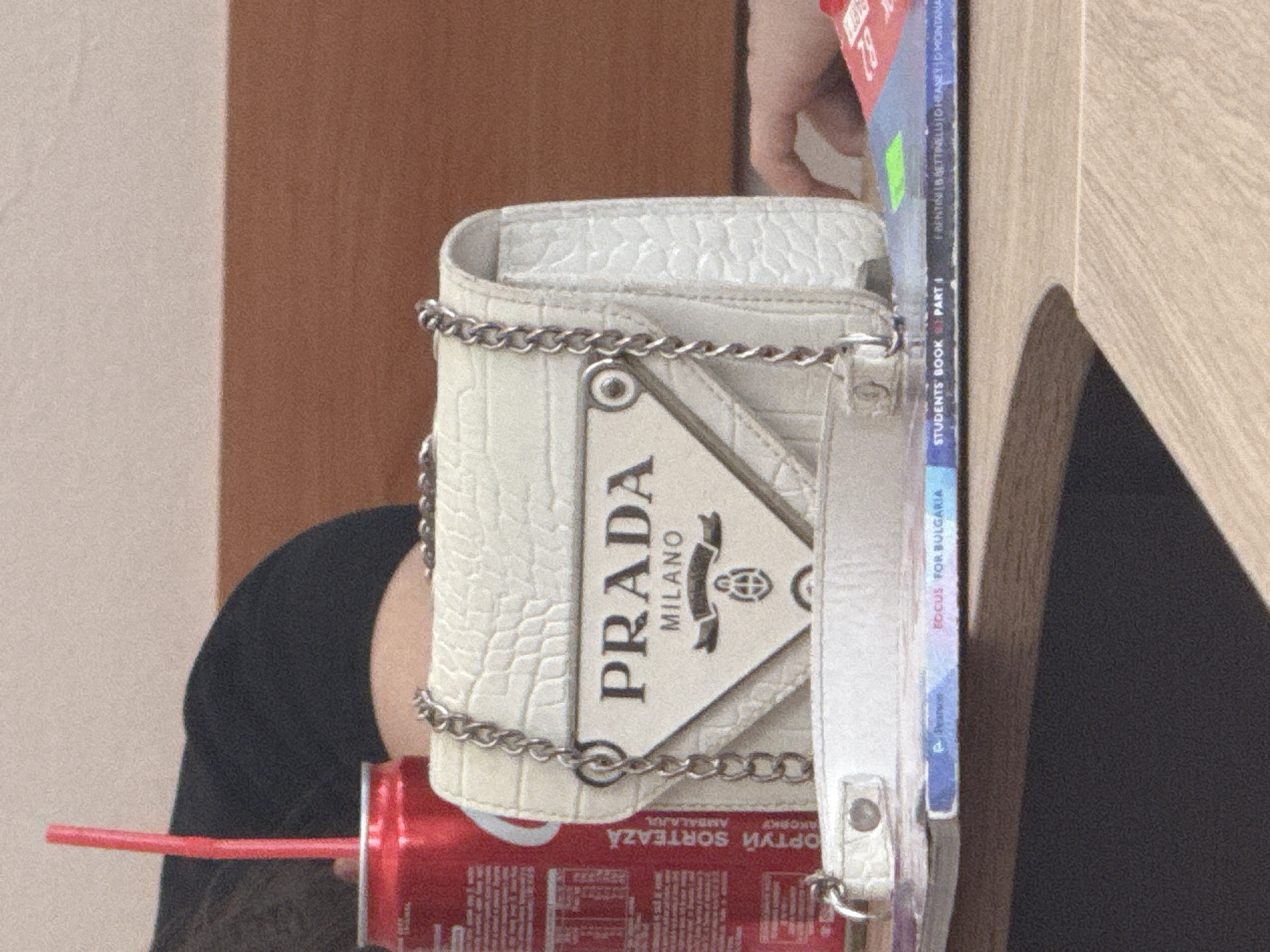The image features a white, textured Prada Milano purse with a silver chain strap resting on top of a red can with a red straw. The can appears to be an “ОРТУЙ SORTEAZĂ” brand soda. Next to the purse and can is a "Focus for Bulgaria" textbook. The text on the spine of the book reads “FRENTINI BRETTINELLI GHEANEYD MONTANA STUDENTS BOOK PART 1." The location is likely Elhovo, Bulgaria given the text on the can and book spine. Other background details include a portion of a wooden table, black fabric, a light brown wall and a partially visible hand.