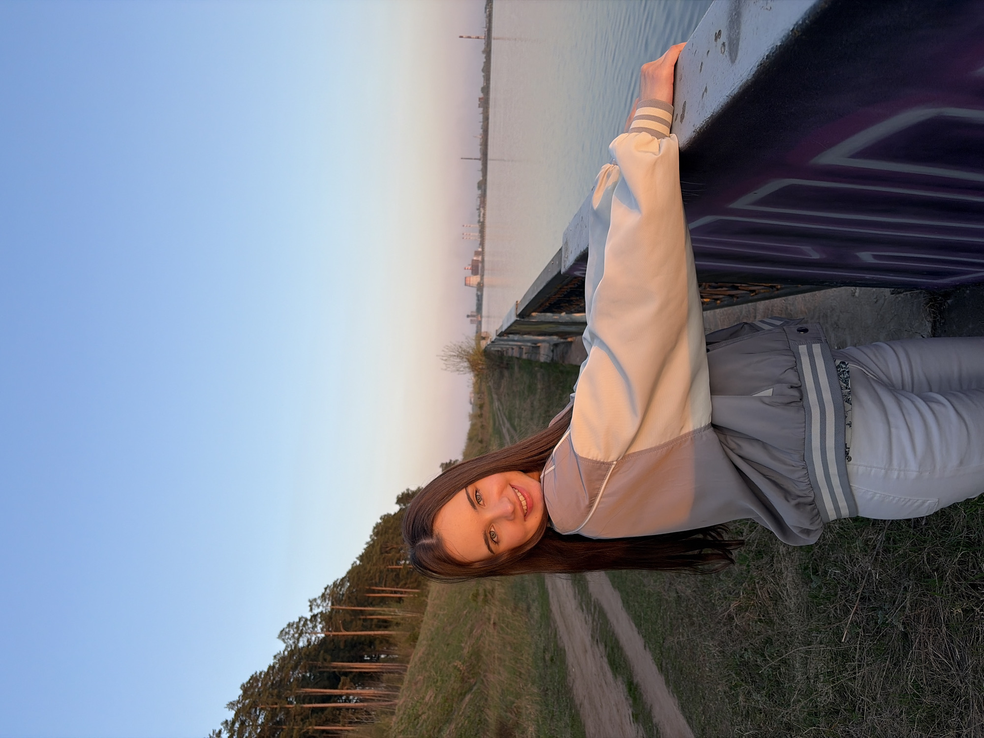 The image depicts a young woman on a bridge in Izhevsk, Russia. She is leaning against the bridge's railing with her hands, looking at the camera and smiling. She is wearing a light-colored jacket with gray accents and matching pants. The bridge railing has graffiti on its underside. In the background, there is a body of water, with buildings and smokestacks visible on the far shore, typical of industrial areas near rivers. The sky transitions from blue to a lighter hue, suggesting either dawn or dusk. Below the bridge, there's a grassy area with trees and a dirt path. The location is identifiable as Izhevsk due to the presence of the iconic Izhevsk dam and the industrial landscape along the shore of the Izhevsk reservoir.