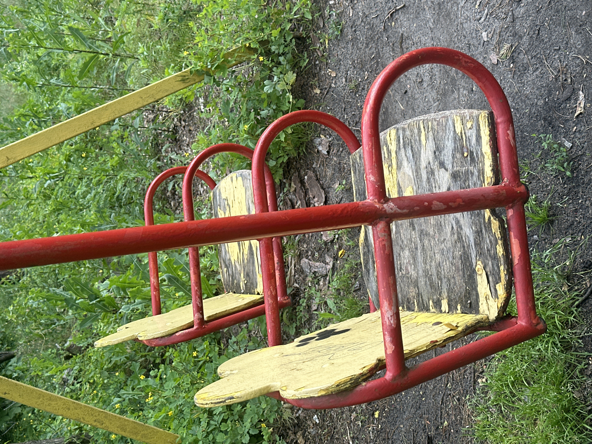 The image shows a close-up, slightly tilted view of a red metal merry-go-round with three yellow wooden seats. The seats are somewhat worn, with peeling yellow paint revealing the gray wood underneath. The merry-go-round is situated outdoors, surrounded by lush green vegetation, including tall grasses and small leafy plants, some with tiny yellow flowers. The ground beneath the merry-go-round is a mix of dark soil, small stones, and scattered dry leaves and pine needles, suggesting a natural, possibly wooded, environment.
The red metal frame of the merry-go-round is prominent, showing some signs of rust and wear, particularly at the joints. A yellow metal bar, likely part of the merry-go-round's structure, is visible in the foreground and extending diagonally across the upper left of the image. The lighting suggests it might be daytime, possibly overcast or in the shade, as there are no harsh shadows. The overall impression is of an old, well-used playground apparatus in a natural setting. No people are visible, and the scene conveys a sense of stillness and perhaps a touch of melancholy due to the visible wear and tear. No text is present in the image.