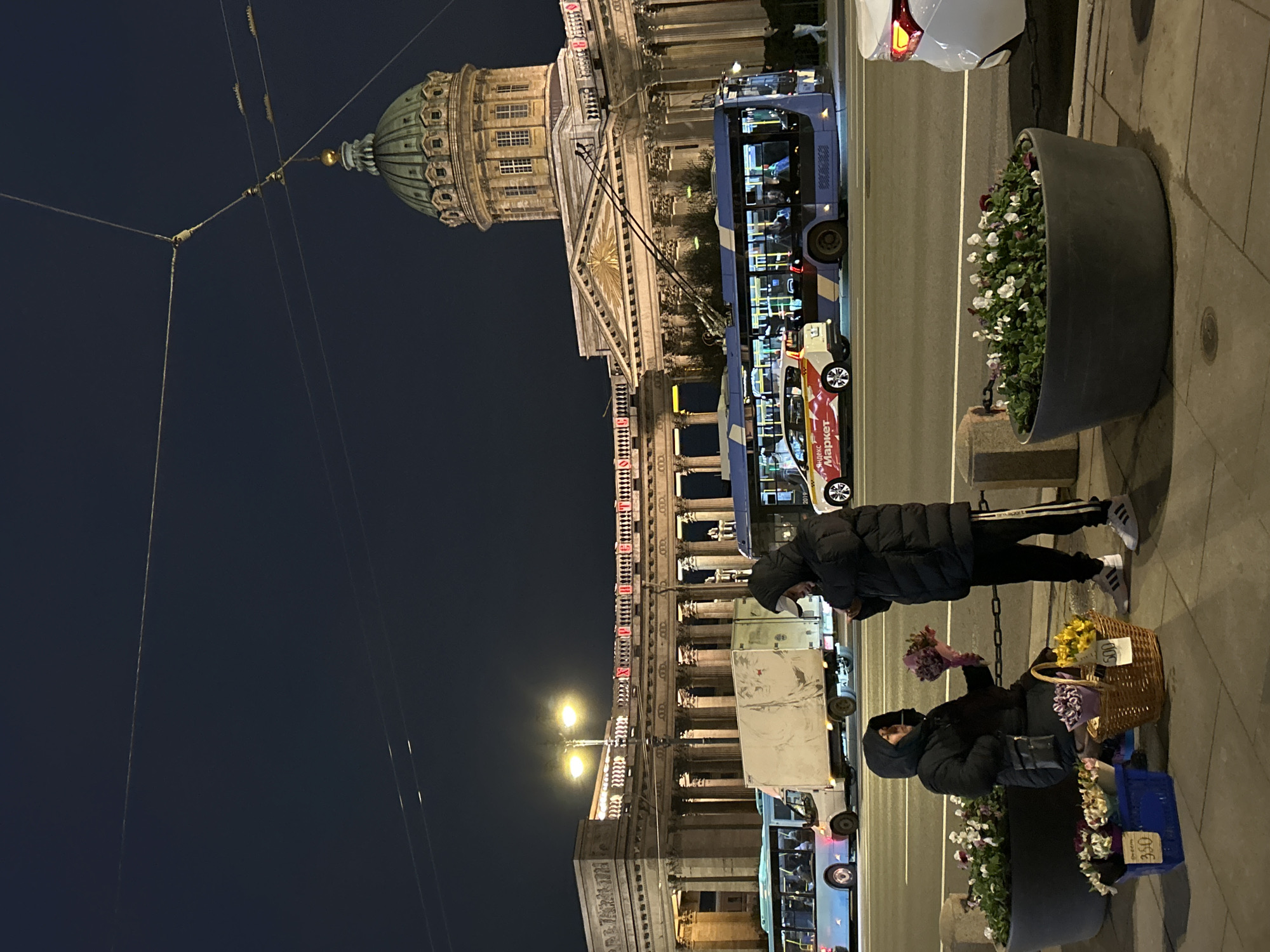 This nighttime photograph captures a bustling street scene in Saint Petersburg, Russia, with the iconic Kazan Cathedral prominent in the background. The sky is dark, indicating late evening or night, with artificial lights illuminating the street and buildings.
In the foreground, two individuals are on the sidewalk. One person, wearing a dark puffer jacket, stands, looking down. The other, similarly dressed in a dark winter coat, is crouched or seated next to several flower arrangements. This person appears to be a flower vendor, holding a bouquet of purple flowers. Various bouquets are displayed in wicker baskets and a blue crate, with price tags of "500" and "350" (likely rubles) visible next to them. Large dark gray planters filled with white and purple flowers line the sidewalk, adding to the urban flora.
The street, partially visible, shows a blue double-decker bus, likely a tourist vehicle, and a yellow taxi with "Яндекс Маркет" (Yandex Market) branding on its side. Other vehicles are also present. Overhead, power lines or tram wires crisscross the dark sky, connecting to poles.
The Kazan Cathedral dominates the background with its distinctive green dome and classical architectural features, including columns. Further down the street, another large building with columns features illuminated red text on its facade. The overall atmosphere is that of an active city evening, with commerce and public transport in operation against a backdrop of historic architecture.