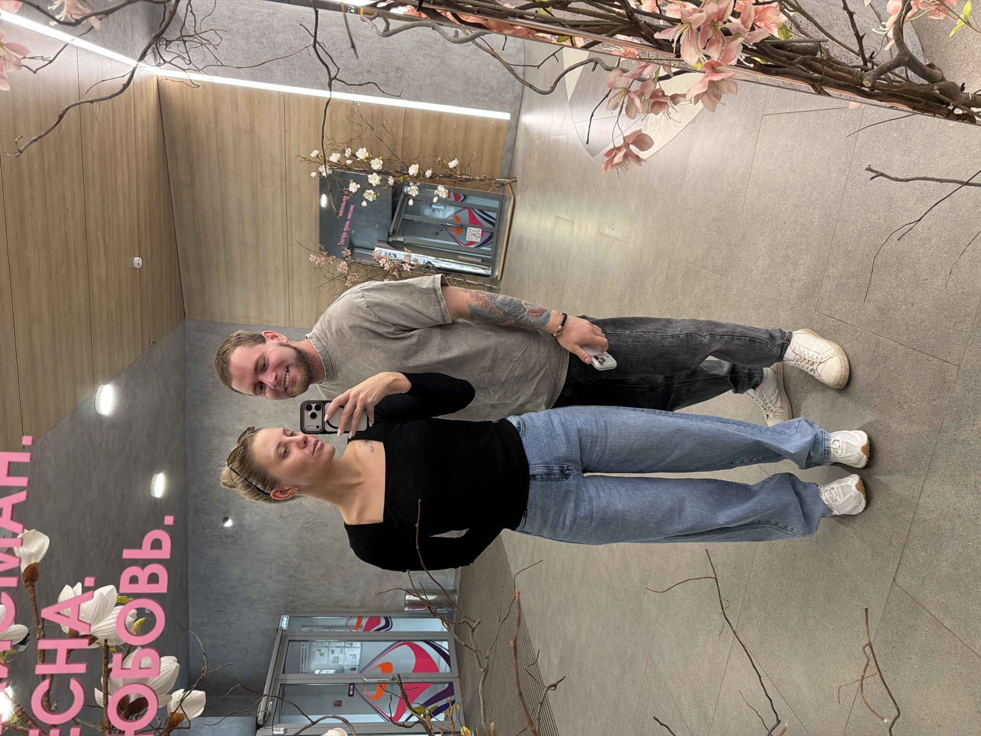 The image shows a reflection of a man and a woman taking a selfie in front of a mirror in a public space. The woman is wearing a black long-sleeved top and blue jeans with white sneakers. The man is wearing a grey t-shirt and dark jeans with white sneakers. He has tattoos on his left arm. They are holding a smartphone to take the picture.
The environment includes interior décor, with artificial trees with pink flowers, and signage in Russian. The location can be identified as Izhevsk, Russia based on the signage "ЧАЙ, КОФЕ, РОМАН". There are shop fronts reflected in the mirror, suggesting a shopping mall or commercial space. The floor is grey tiled.