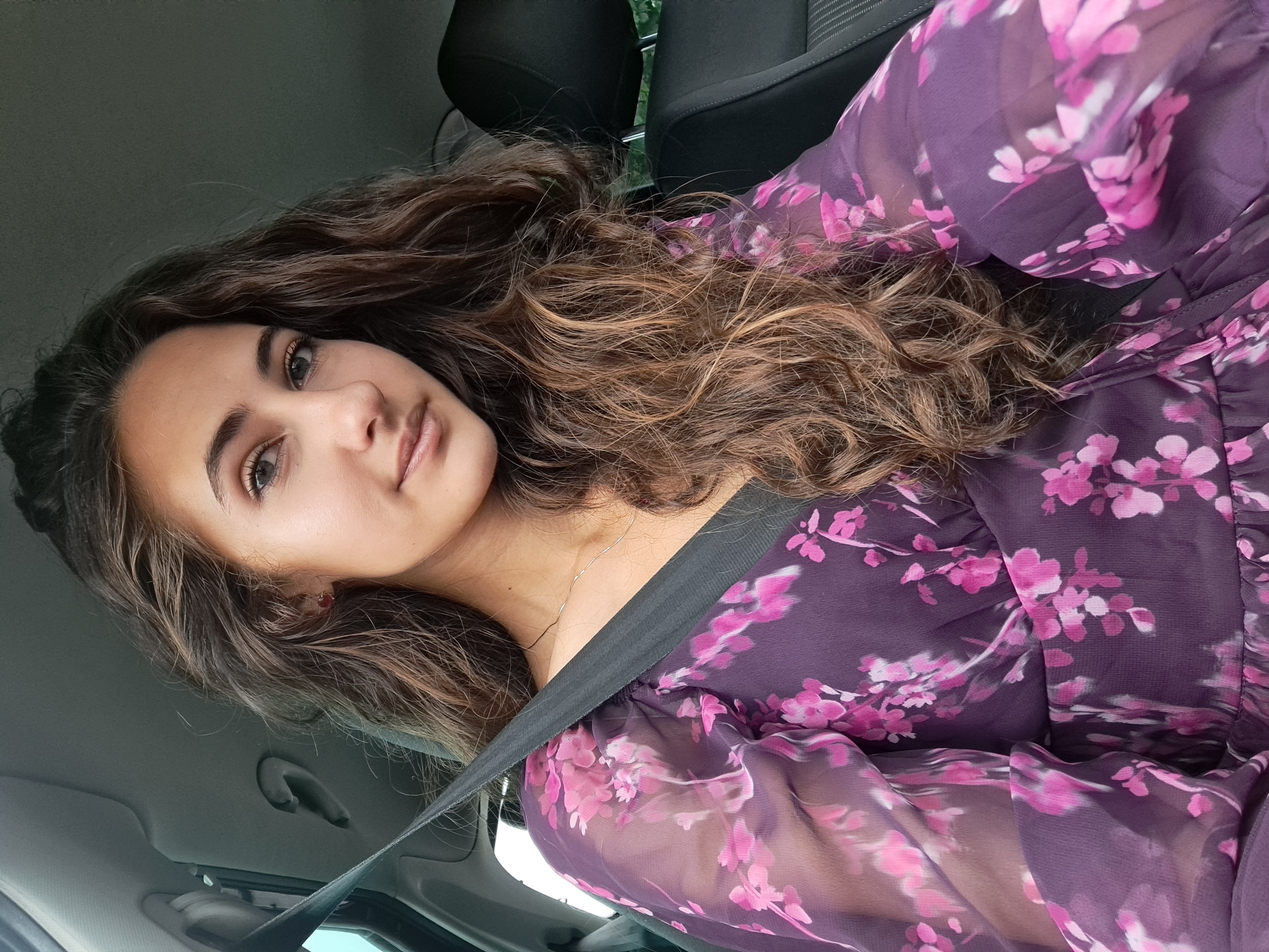 A young woman is captured in a portrait-style photograph taken inside a car. Her head is slightly tilted, and she looks directly at the camera with a subtle, serene expression. She has long, wavy brown hair with lighter highlights, which frames her face and cascades over her shoulders. Her eyes are brown, and she appears to have natural makeup.
She is wearing a sheer, deep purple top or dress featuring vibrant pink floral patterns. A black seatbelt diagonally crosses her chest, indicating she is seated in a moving or parked vehicle. A delicate silver necklace is visible around her neck.
The background shows elements of the car's interior, including dark grey or black car seats with a textured fabric and a light grey headliner. Through the windows, faint hints of what appears to be greenery or the sky are visible, suggesting the photo was taken during the daytime. No text is visible in the image.