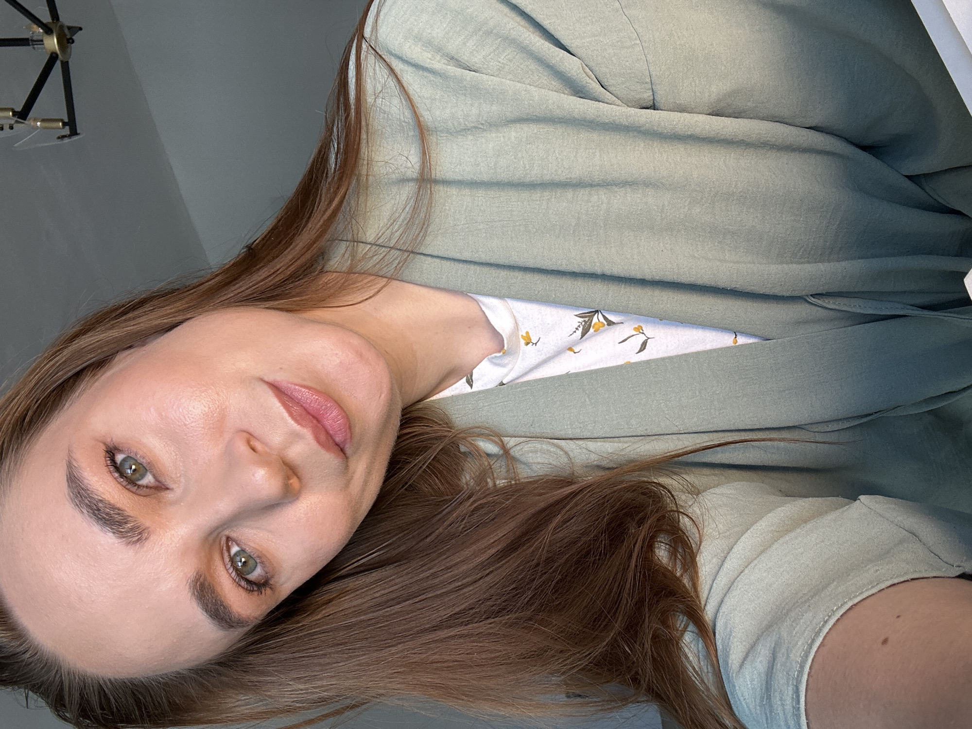 A woman with light skin, green eyes, and brown hair is posing for a selfie indoors. She is wearing a light green garment over a patterned top. A modern light fixture is visible in the background. The location in the city of Kharnavy, Russia cannot be determined from the image.