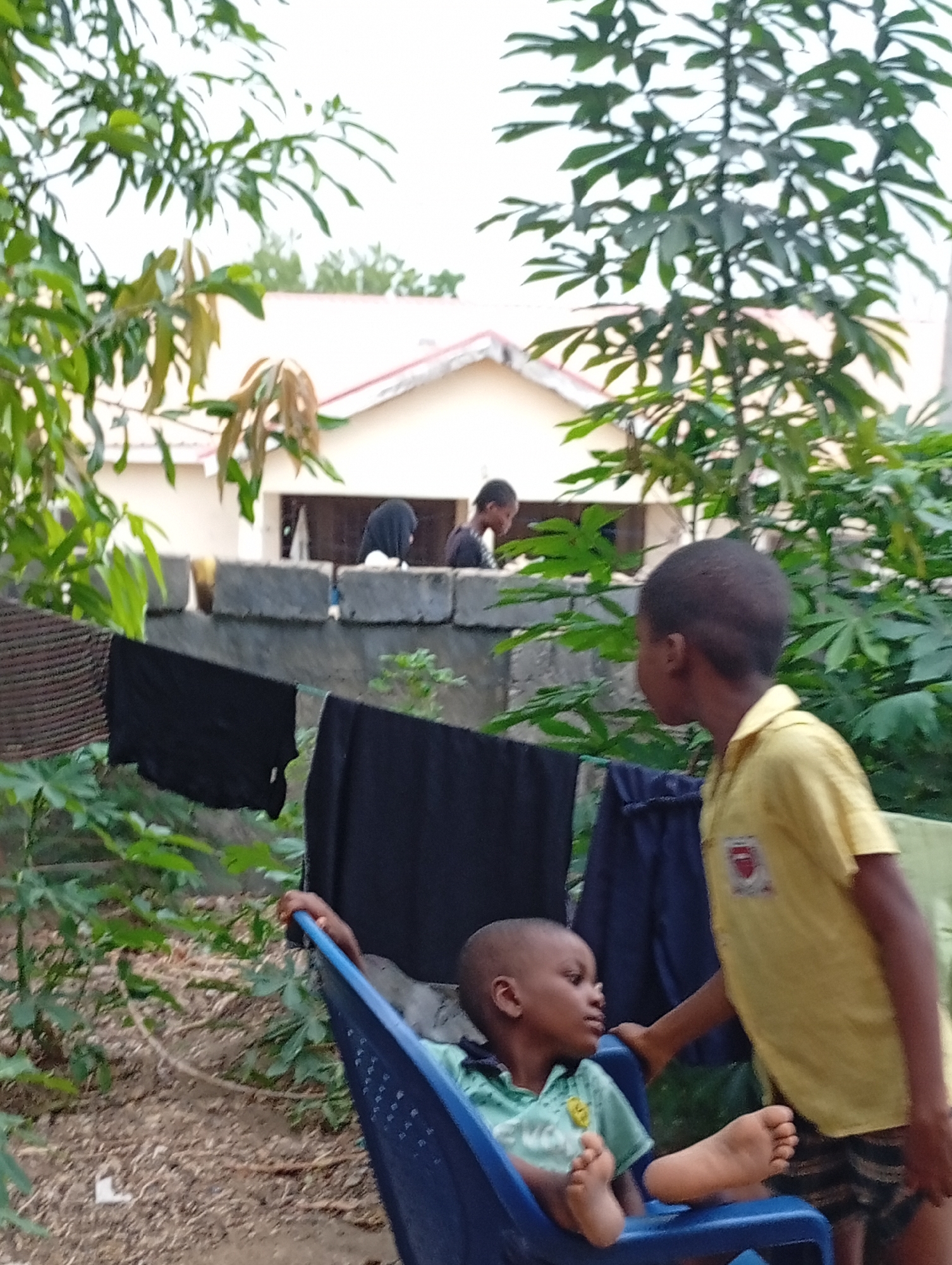 The image depicts two children in an outdoor setting, potentially a backyard or yard area. One child is sitting in a blue plastic chair with their feet extended, wearing a light green collared shirt. The second child is standing next to the chair, wearing a yellow shirt and patterned shorts. Clothes are hanging on a clothesline in the background, with black and blue items visible. Behind the clothesline, there is a concrete structure that could be a wall or the base of a building. In the background, there is a building with a white exterior and a red-tiled roof. Other people are visible standing behind the concrete structure near the building. Trees and vegetation surround the scene. The image appears to be taken during the daytime. The location is in Adankolo, Nigeria.