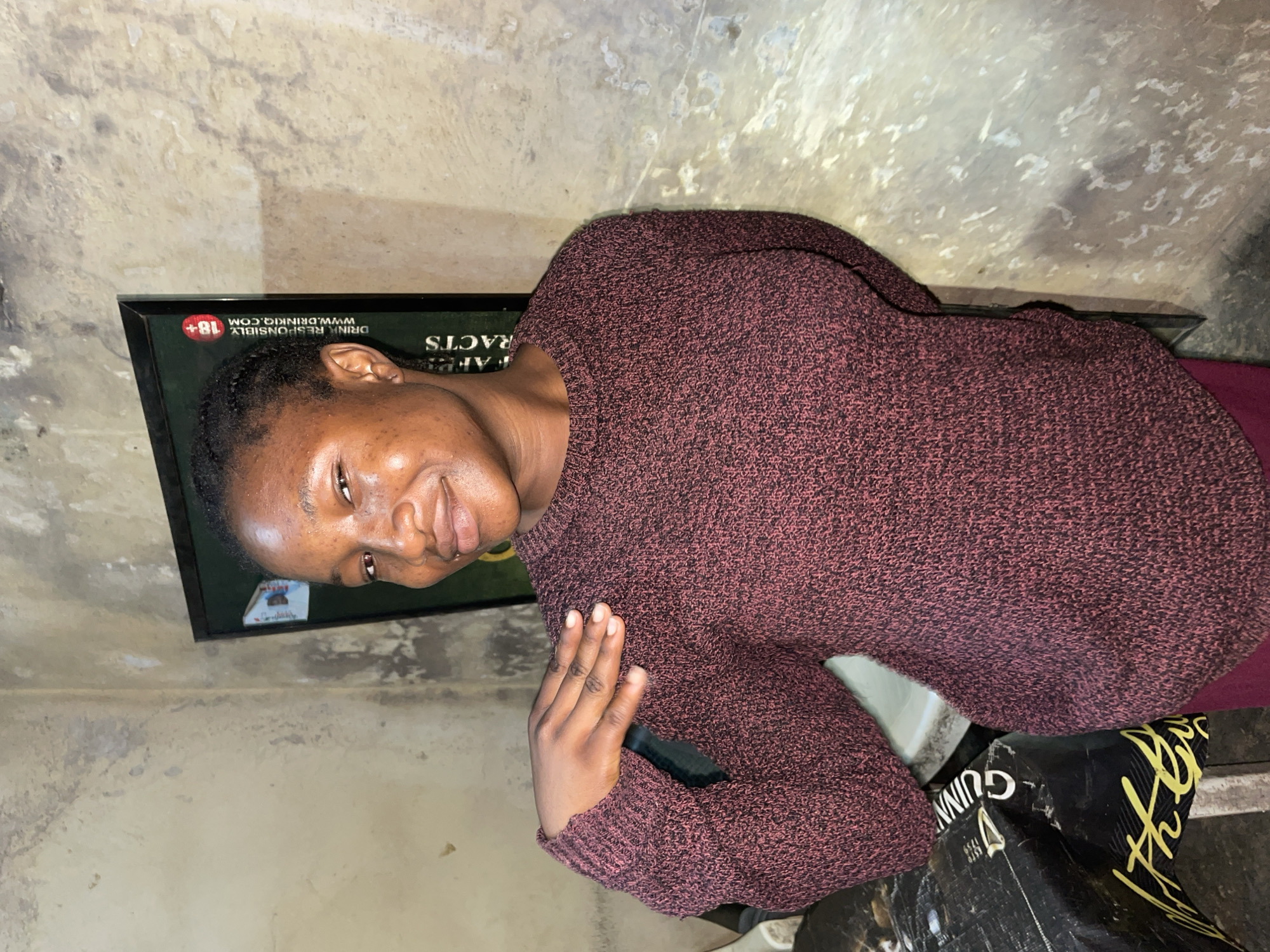 A person with dark skin and short, dark hair is oriented sideways in the image. They are wearing a dark maroon sweater. Behind them is a framed advertisement with the words "DRINK RESPONSIBLY" and "WWW.DRINKIQ.COM," with "+18" in a red circle. The wall behind the frame is off-white with mottled staining. The person's right hand is visible, holding what appears to be a black smartphone. Below the person is a black box with yellow lettering, "GUINN." The environment appears to be indoors. Jos, Nigeria cannot be determined from the given image alone.