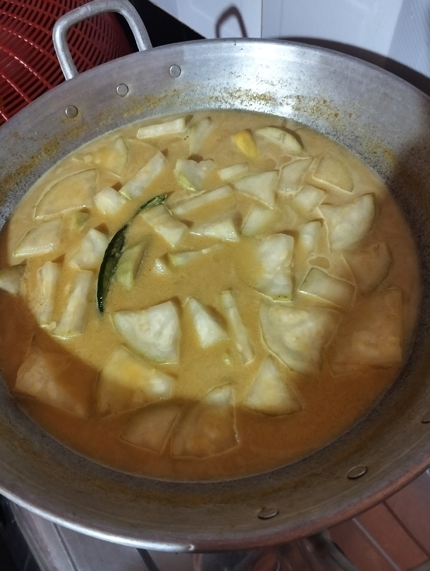 A close-up, overhead shot shows a large, round, aluminum cooking pot filled with a light orange, creamy curry. Inside the curry are numerous pieces of light-colored, sliced vegetables, appearing to be winter melon or a similar gourd. One dark green chili pepper is visible, submerged in the curry. The pot has two metal handles and is situated on a stovetop. The background is blurred, but suggests a kitchen environment with a red woven object to the left and a tiled wall behind. There are no people visible in the image. The scene is of food being cooked in a home kitchen, possibly in Payagaboh, Indonesia.