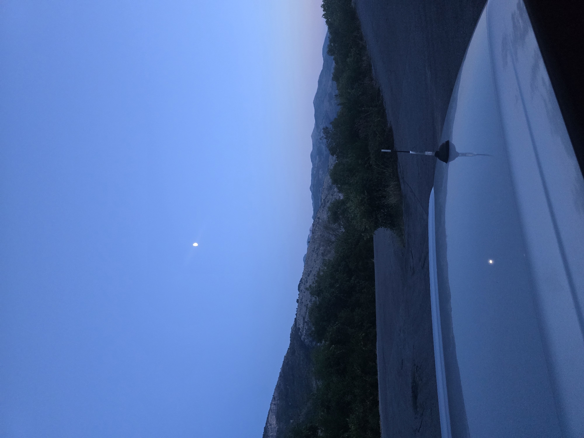 The image depicts a twilight scene with a clear, deep blue sky. A small, bright crescent moon is visible in the upper portion of the sky. The landscape consists of rolling hills covered in dark green foliage and scrub, transitioning to a lighter, more arid slope with sparse vegetation in the background. In the foreground, the hood of a silver-colored vehicle is prominently featured, reflecting the sky and a small point of light, likely the moon. A black car antenna with a transparent tip protrudes from the hood. A paved road curves through the scene, disappearing into the shadows of the hills. The overall impression is one of a serene, natural landscape at dusk. The location is Klopot, Montenegro.