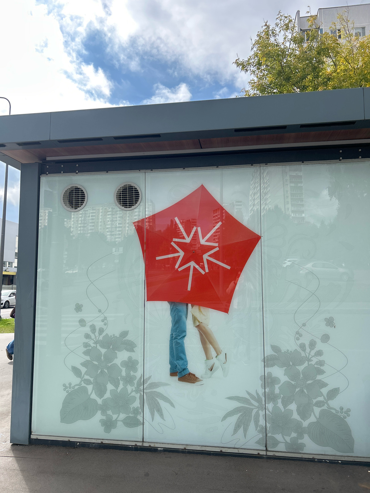 The image shows a bus stop shelter with a large advertisement on the glass panels. The advertisement features a graphic of a red umbrella with white arrows pointing outwards from its center. Partially visible beneath the umbrella are the legs of two people, one wearing blue jeans and brown shoes, the other wearing white sneakers. The background of the advertisement is a light translucent surface with subtle floral patterns. Through the glass of the shelter, several high-rise apartment buildings are visible, along with trees and a cloudy blue sky. The bus stop itself is made of gray metal and glass. The location is identified as Moscow, Russia, and the image appears to have been taken during the daytime, likely in the autumn given the foliage on the trees.