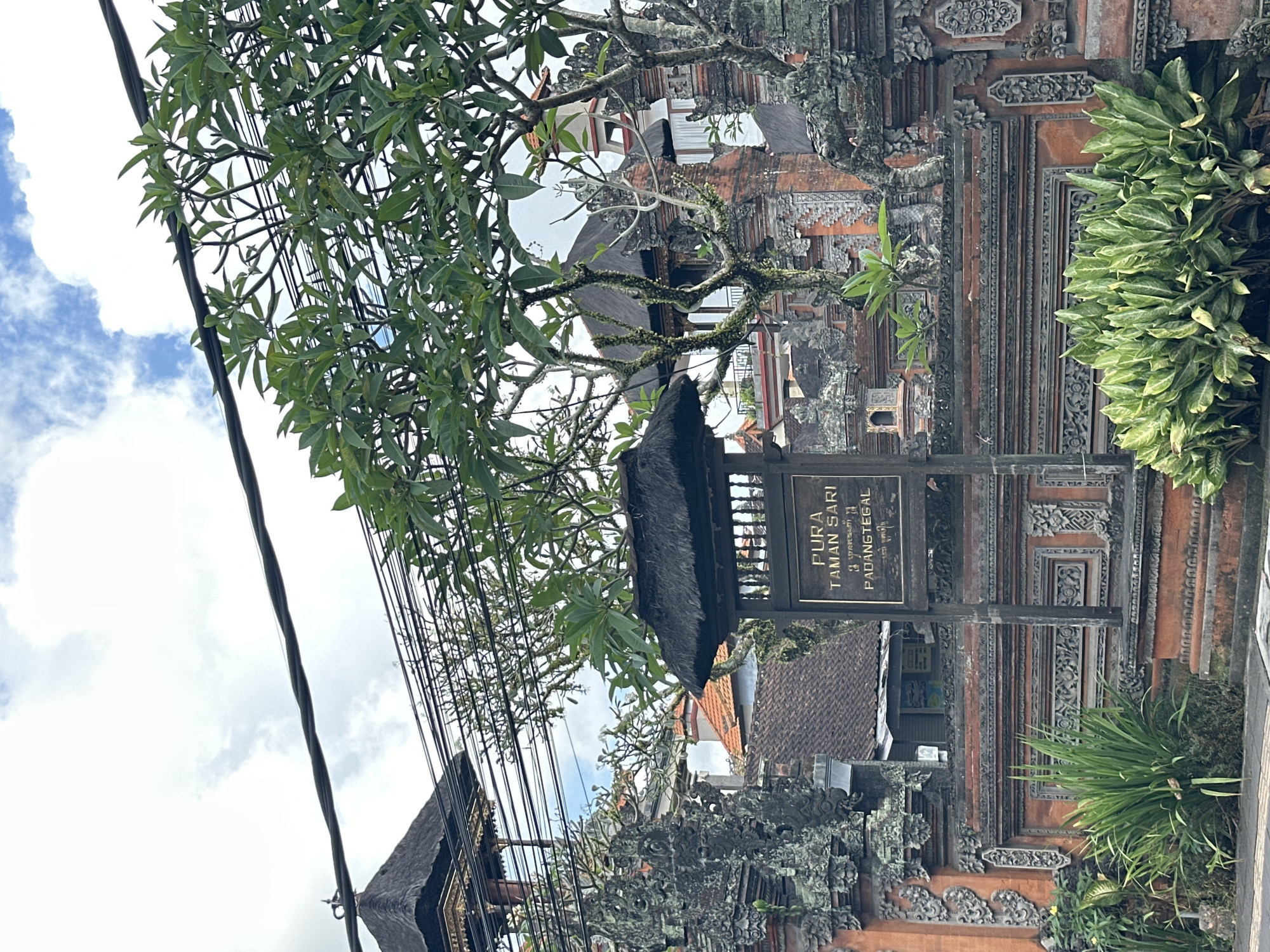 The image shows a sign for "PURA TAMAN SARI PADANGTEGAL" in Ubud, Indonesia. The sign is made of dark wood with gold lettering and is mounted on a wooden post with a thatched roof. The sign is set against a backdrop of traditional Balinese architecture, characterized by intricate stone carvings and terracotta roofs. Lush green foliage, including trees and plants, frames the scene. Power lines are visible across the top of the image, suggesting an urban or semi-urban setting within Ubud. The sky is partly cloudy.