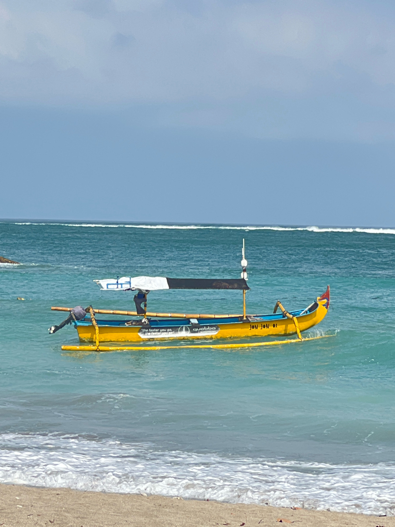 In Kuta, Indonesia, a yellow and blue fishing boat with outriggers floats on turquoise water under a partly cloudy sky. A person in a gray shirt and dark pants is seated on the boat, facing away from the camera. Another person, also in dark clothing, is standing on the boat, near the stern. The boat has a white and black canopy supported by bamboo poles. There are white text markings on the side of the boat, including "JON JON 01" and "the faster you go, the faster you go." In the distance, white waves break on the horizon. The foreground shows a sandy beach with small debris.