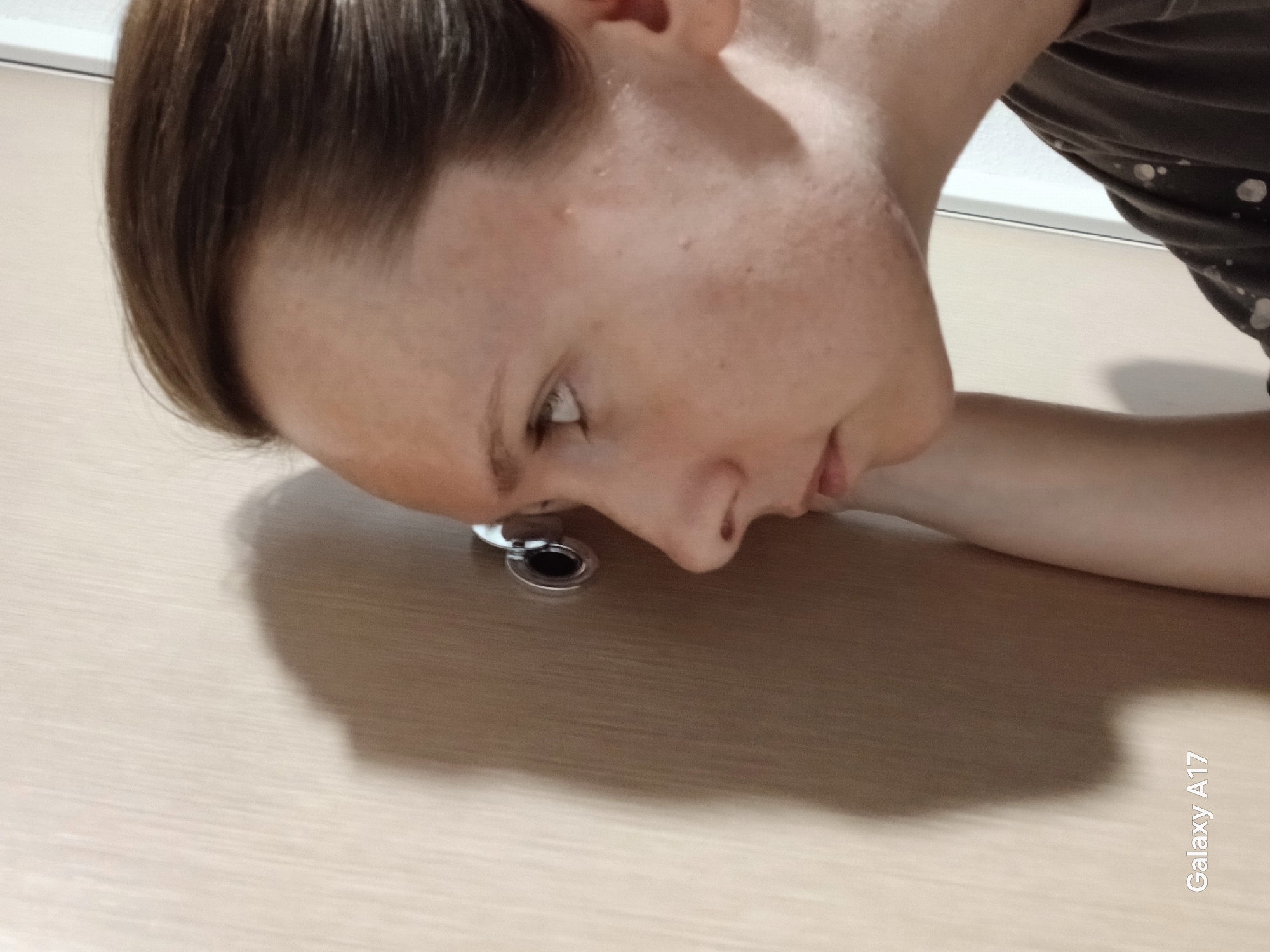 A fair-skinned person is bending down, looking through a peephole in a light-colored door. The person has short brown hair, and their nose is close to the peephole. The person's right arm rests on the door surface. The location of the image is unknown but it is not Yekaterinburg, Russia. "Galaxy A17" is visible in the bottom right corner of the image.