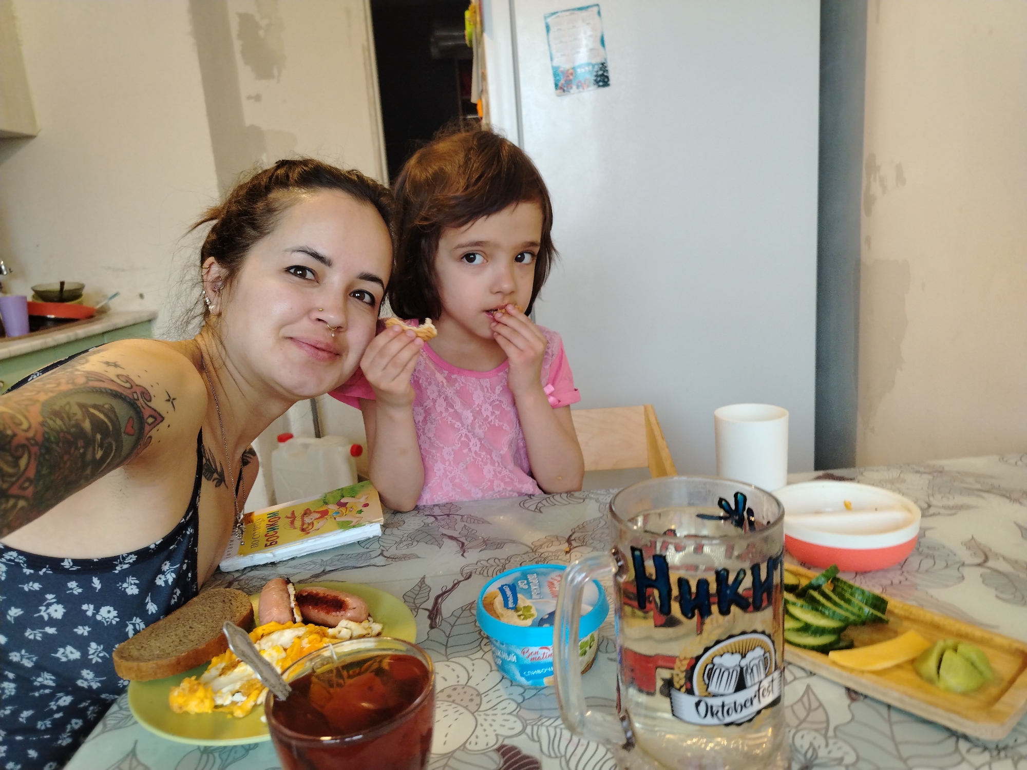 The image shows a woman and a child at a table with food. The woman has dark hair pulled back, a nose ring, and tattoos on her arm. She is wearing a dark floral-patterned top. The child has brown hair and is wearing a pink shirt. They are sitting at a table covered with a patterned tablecloth. The table is set with a plate of eggs, sausage, and bread, a glass of dark liquid, a container of what appears to be dessert, and a mug with the word "Ники" (Niki) and "Oktoberfest" on it, possibly referring to a local tradition or event. There is also a wooden tray with sliced cucumbers and cheese. In the background is a refrigerator and a wall with peeling paint. Given the context, this appears to be a casual meal at home. There is no explicit indication the image was taken in Noviy, Russia.