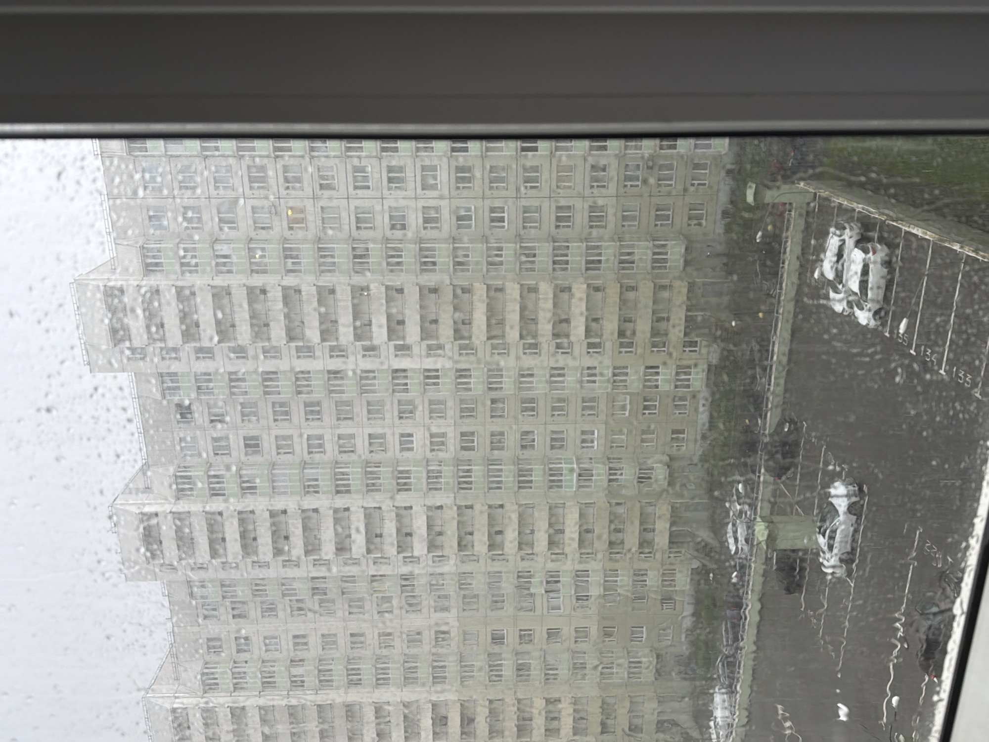 This is a medium-shot, eye-level, vertical photograph taken through a rain-streaked window, looking out onto a large, multi-story residential building and an adjacent parking lot. The building is a typical Soviet-era panel building, grey and uniform, with rows of identical windows and balconies. The sky is overcast, suggesting a dreary, possibly cold, day in Saint Petersburg, Russia.
In the foreground, the window is covered in numerous raindrops, some large and distinct, others forming streaks that distort the view. The reflection of the camera or viewer is visible on the glass.
The parking lot below is wet, indicating recent or ongoing rain. Several cars are parked in designated spots. In the center of the parking lot, a white car is prominently visible, appearing slightly damaged or distressed. To the right of this car, a group of three white SUVs are parked closely together. Further back, a line of cars, mostly dark-colored, are parked along the building's perimeter. The parking lot is sectioned by white lines, some of which are partially obscured by water.
The overall impression is one of a typical urban scene in Saint Petersburg during inclement weather, with the focus on the residential architecture and the mundane details of daily life in a courtyard.
