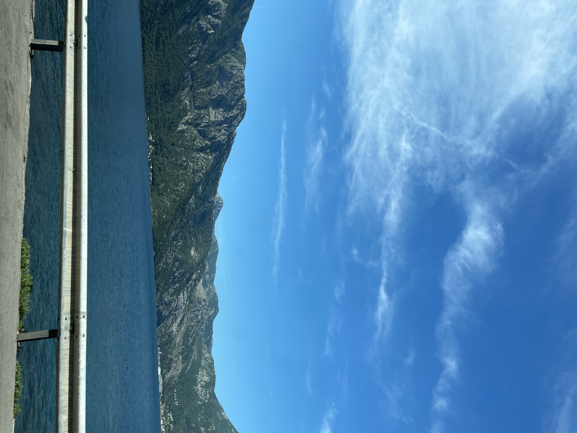 The image captures a scene in Kostanjica, Montenegro, showcasing a view of the Bay of Kotor. A portion of a roadside barrier is visible on the left. Beyond the barrier, there is a body of water, likely a part of the bay. On the opposite side of the water, a mountain range rises prominently, its slopes covered with vegetation. The sky is a clear blue with scattered cirrus clouds.
